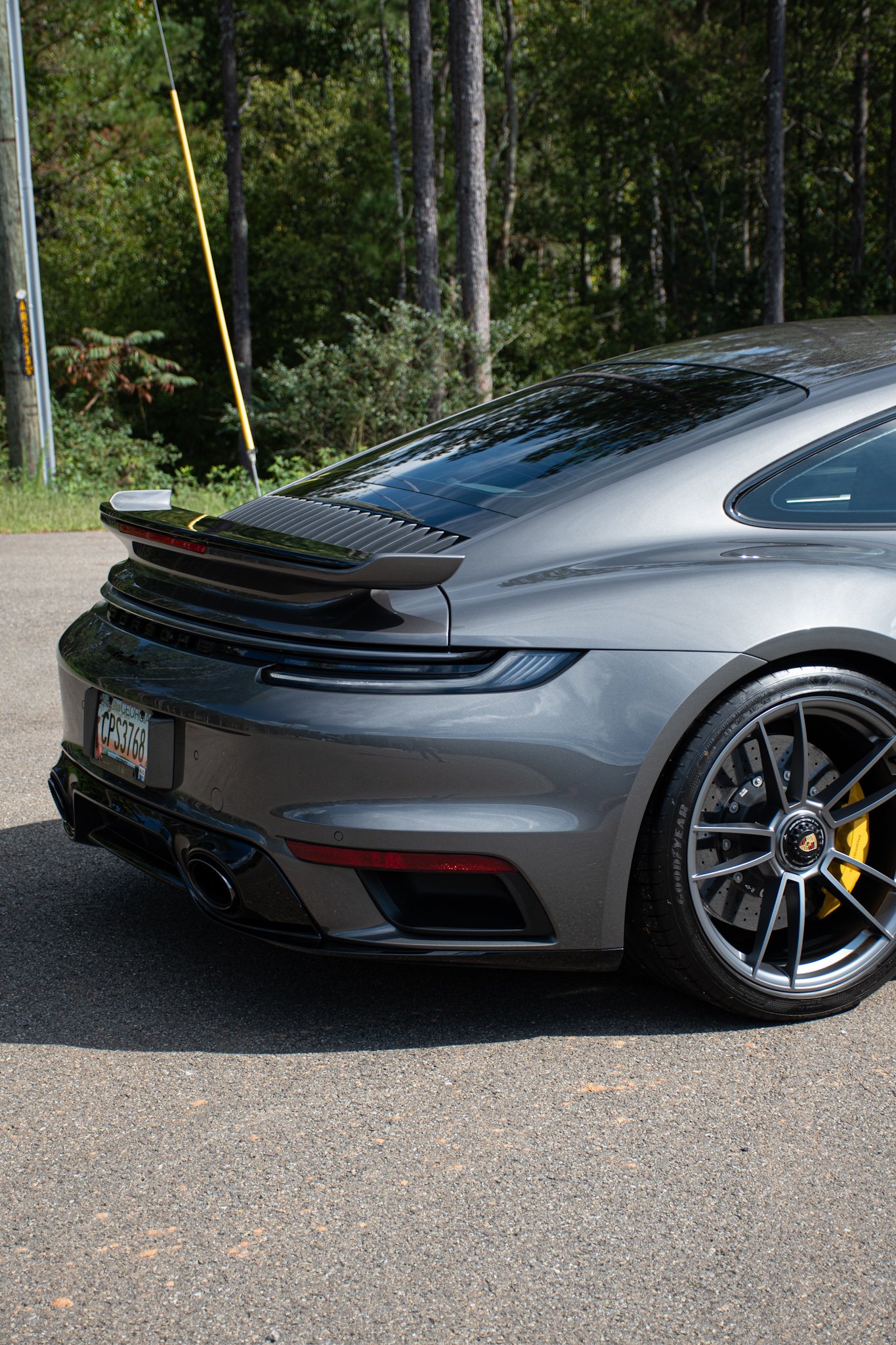 Close-up of the rear side of a gray Porsche sports car parked on a street with a forest background.