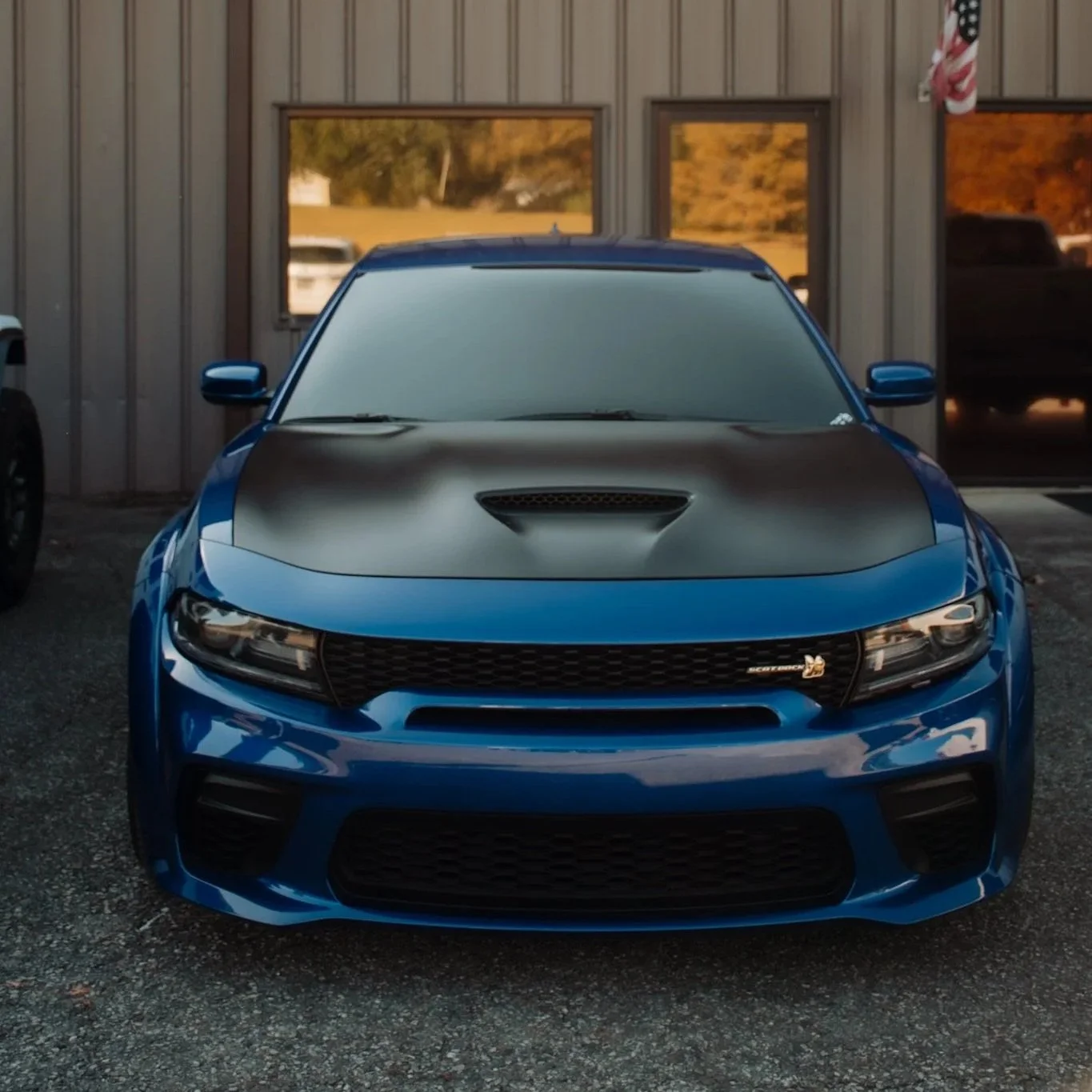 Front view of a blue car with a matte black hood parked outside a building with windows and an American flag.