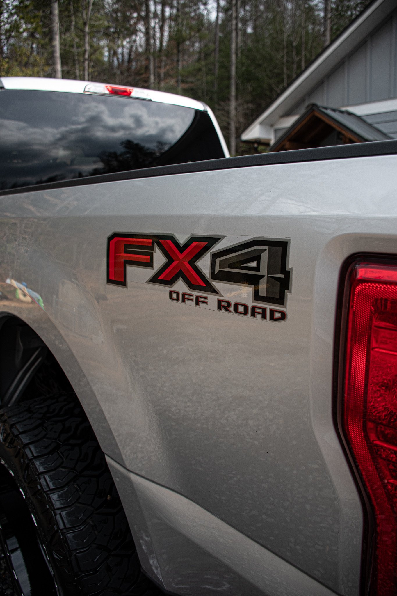 Close-up of a silver pickup truck's rear side with a red and black vehicle model badge reading 'FX4 OFF ROAD' and part of the taillight, with a roof and trees in the background.