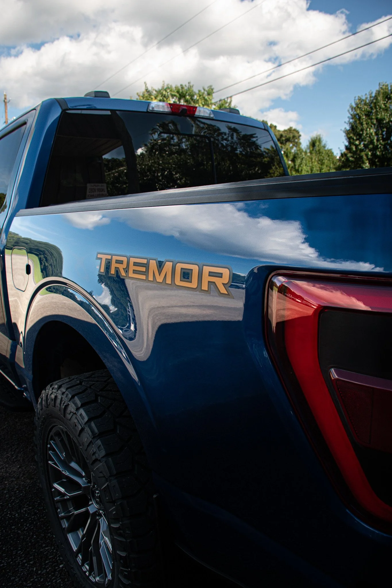 Close-up of the rear side of a blue pickup truck with the word 'TREMOR' visible on the side, chrome accents, large all-terrain tires, and the sky with clouds and power lines reflected on the truck's surface.
