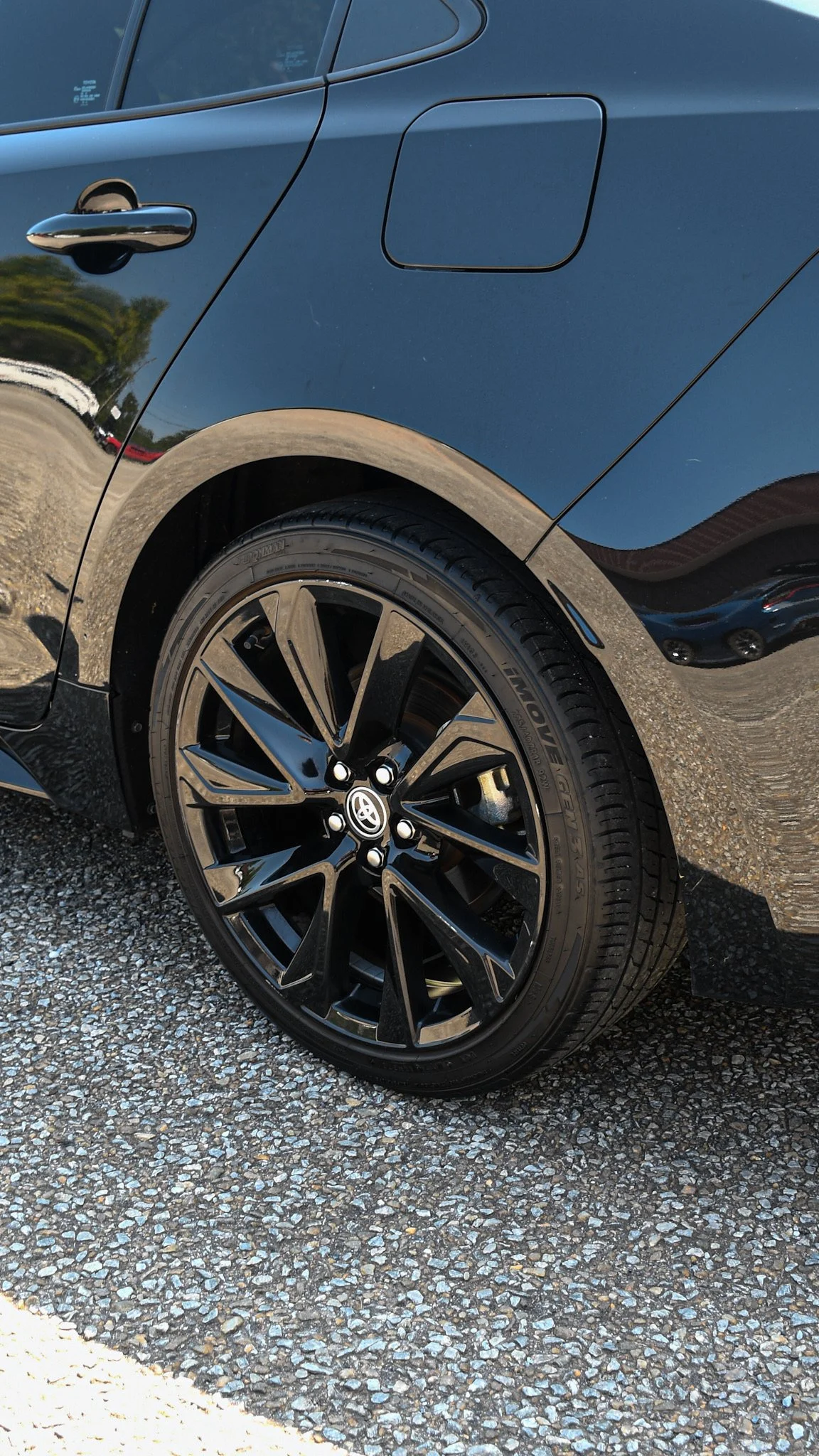 Close-up of a black car's rear side with a shiny rim, tire, and a closed fuel cap, parked on a gravel surface, some trees and other vehicles reflected on the car's surface.