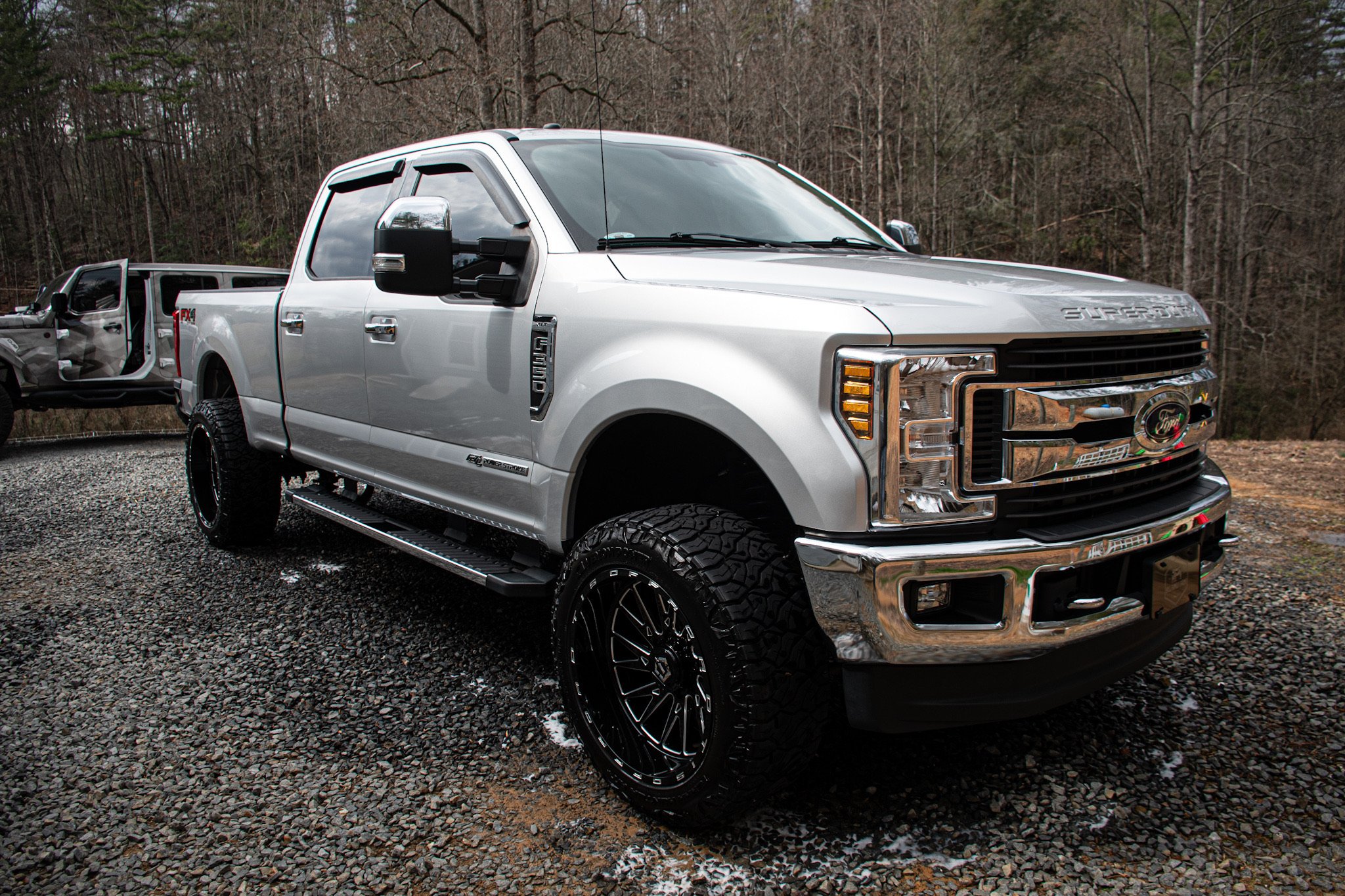 A silver Ford Super Duty pickup truck parked on a gravel surface in front of leafless trees, with its driver's door open.