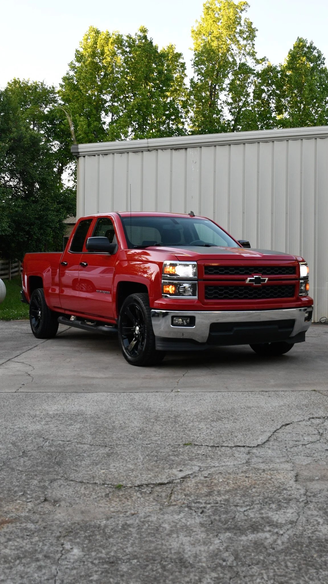 A red Chevrolet Silverado pickup truck parked on a concrete driveway with a white metal building and green trees in the background.