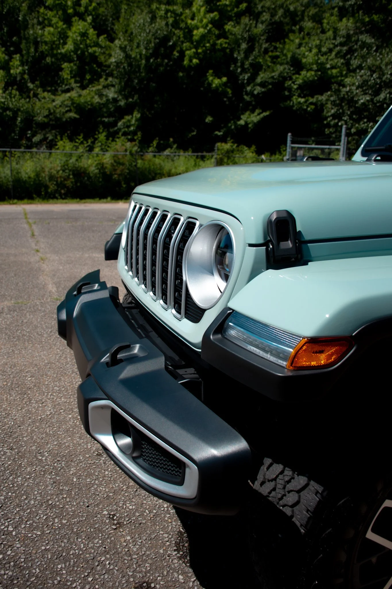 Close-up of the front of a green Jeep SUV parked on a gravel lot with a wooded background.