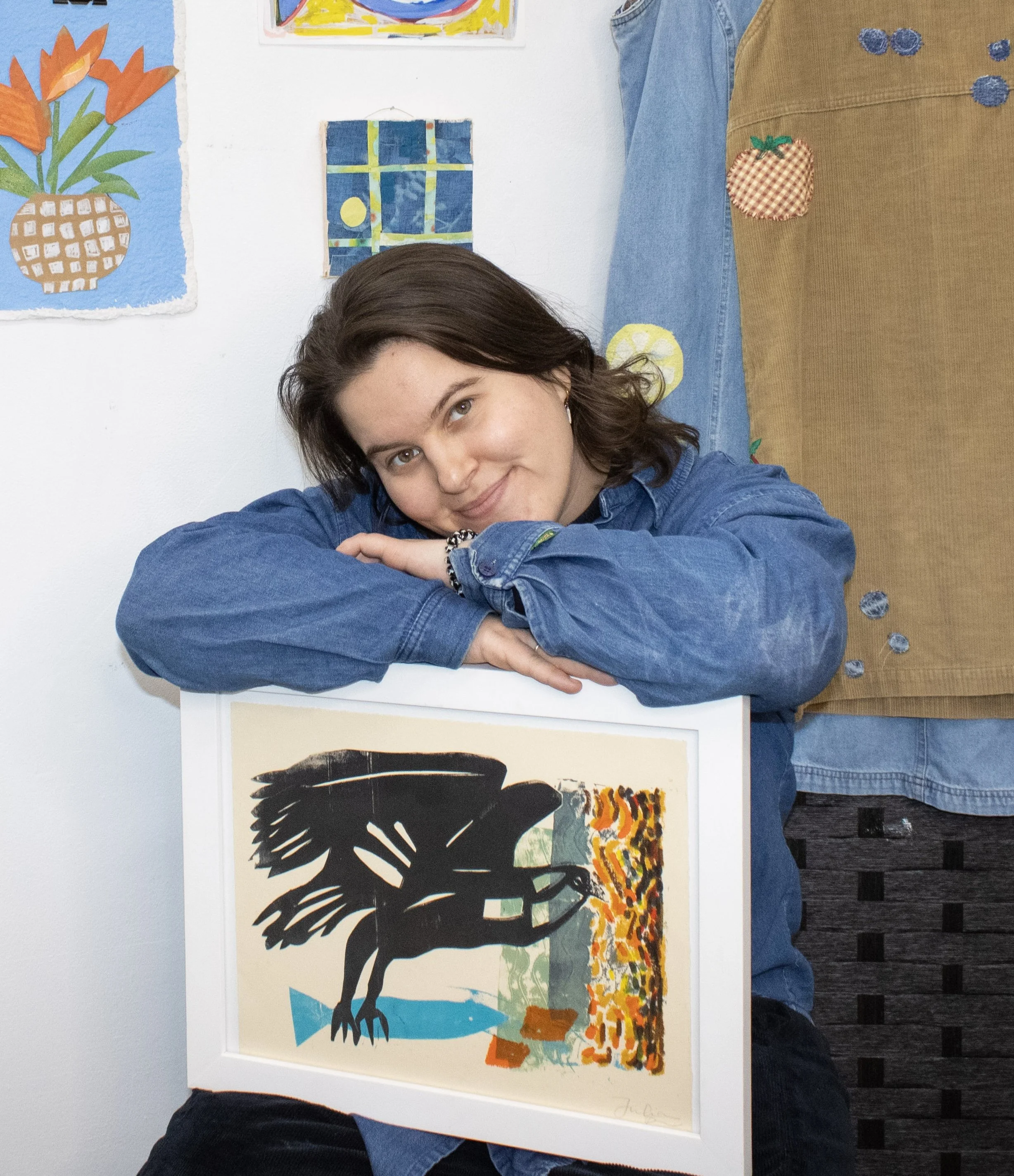 Julia Kagan sitting with her arms crossed resting on a framed picture of her artwork. Julia is a white, femme person with brown hair. Numerous pieces of artwork surround her.