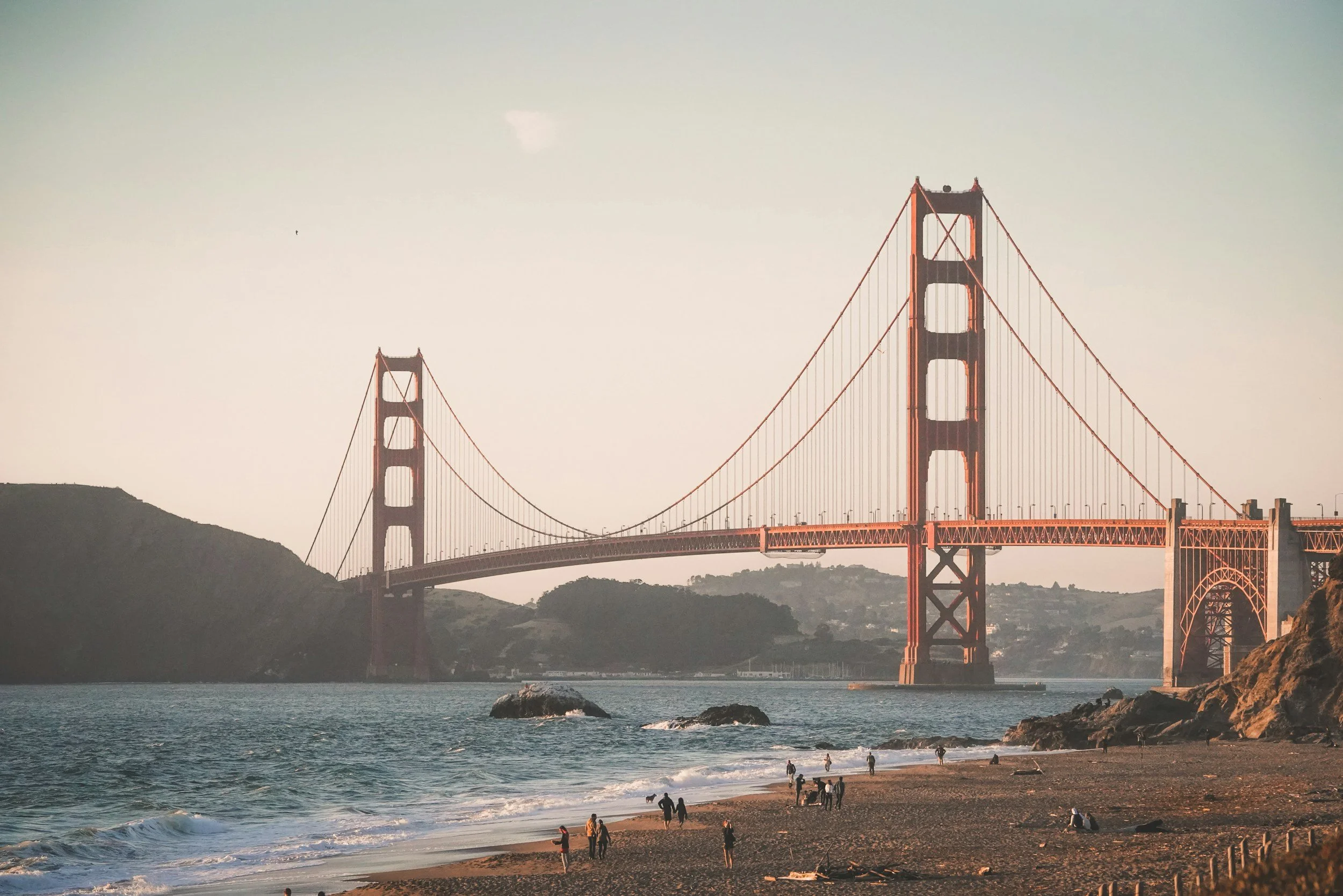 Golden Gate Bridge over water with a sandy beach and people walking along the shore in the foreground, hills in the background, under a clear sky.