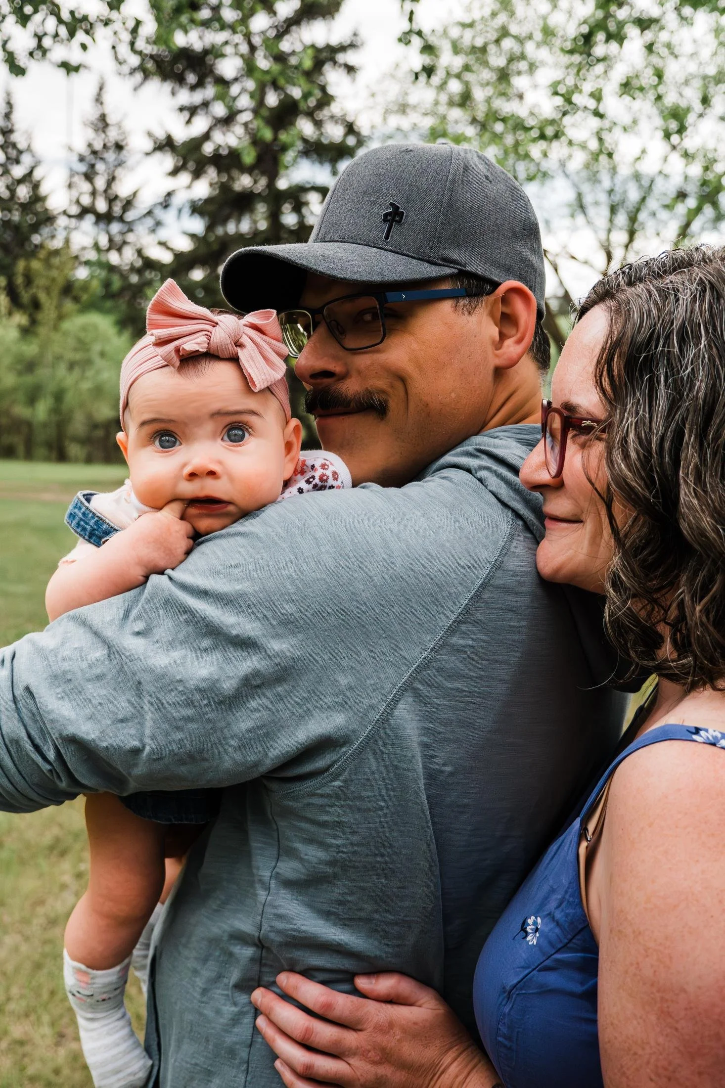 Parents cuddling their baby outdoors, with the baby wearing a pink bow and looking toward the camera.
