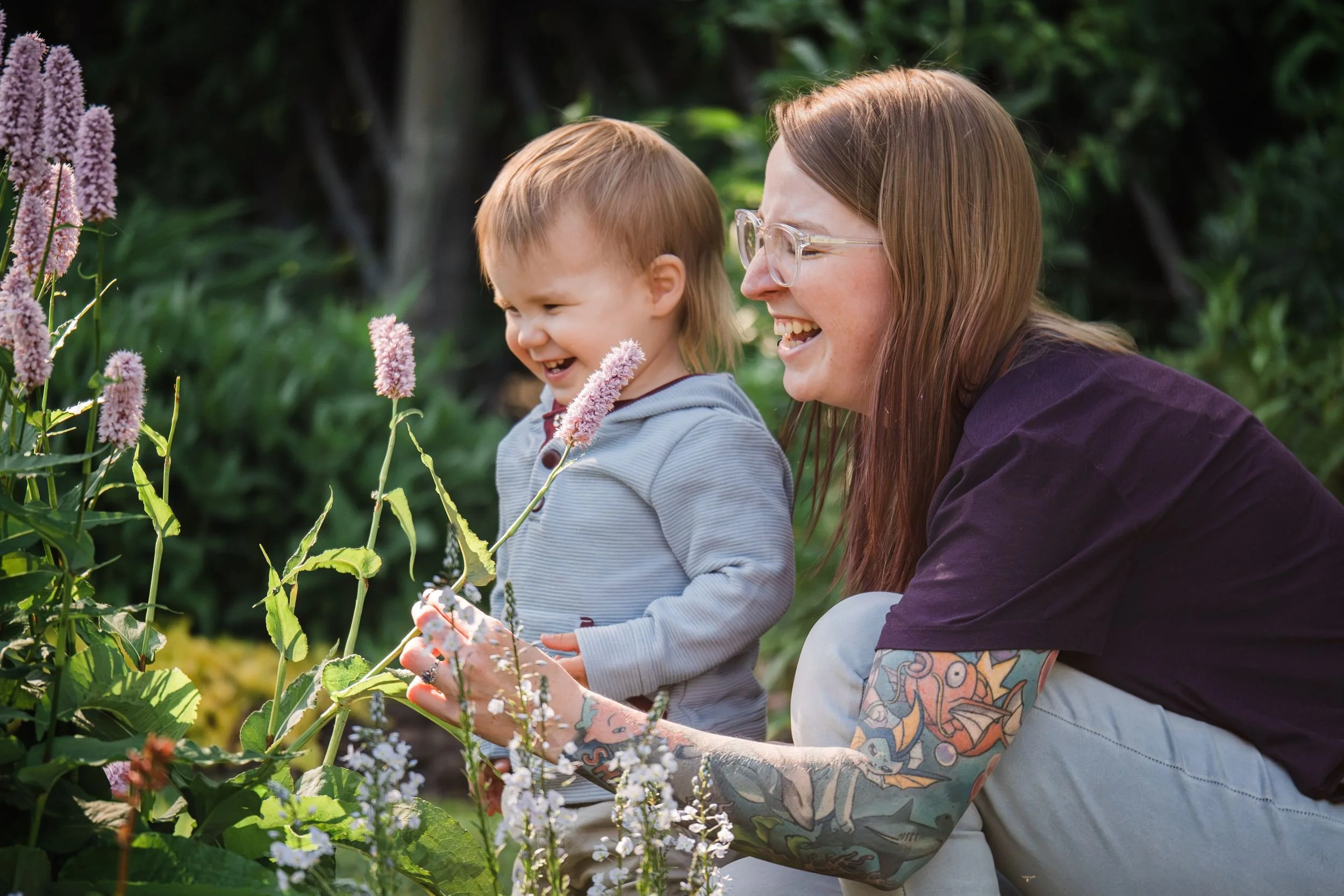 Mother and young child laughing together beside pink and purple flowers in a sunlit garden.