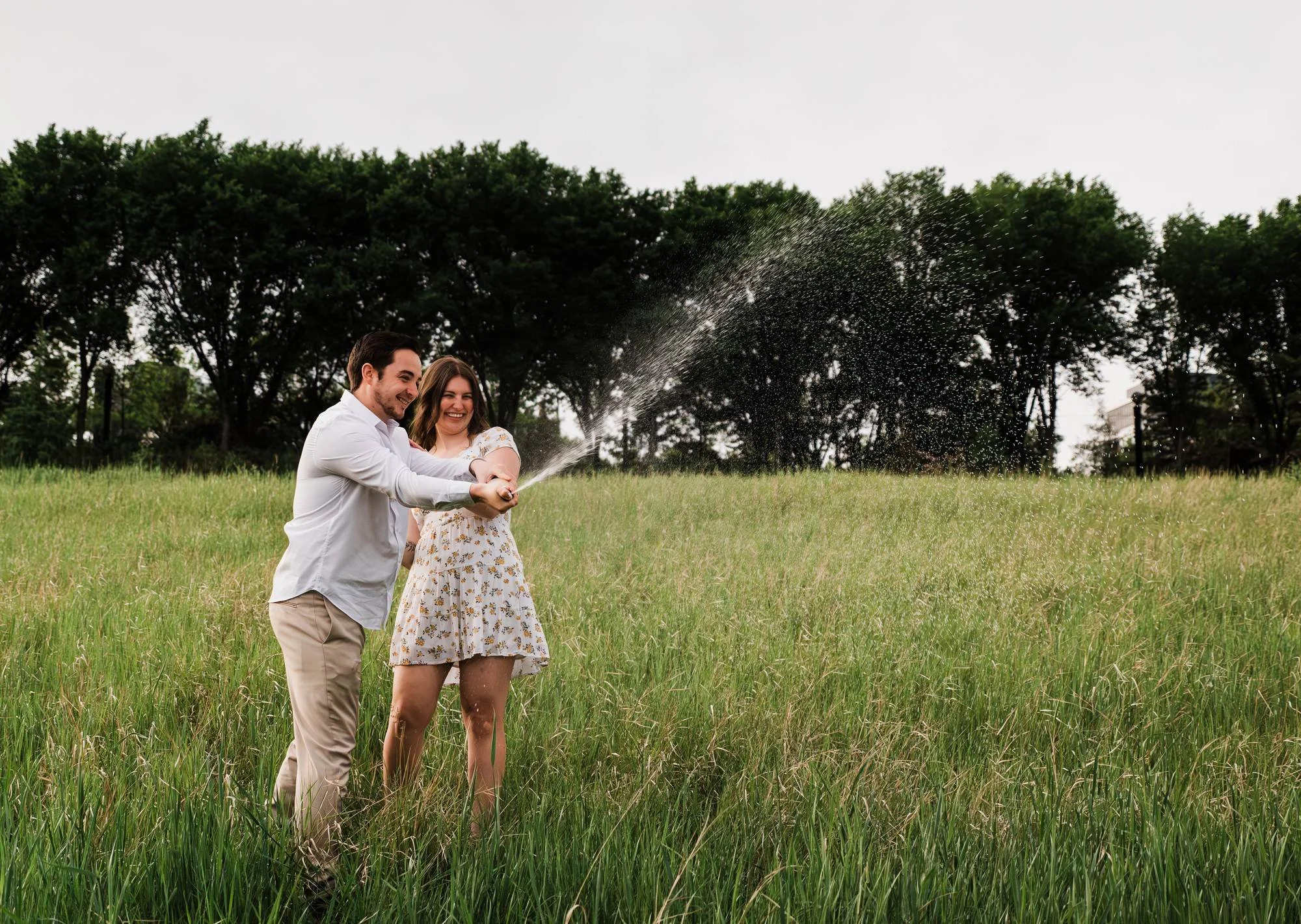 Couple smiling as they spray champagne together in a grassy field during their Edmonton engagement session.