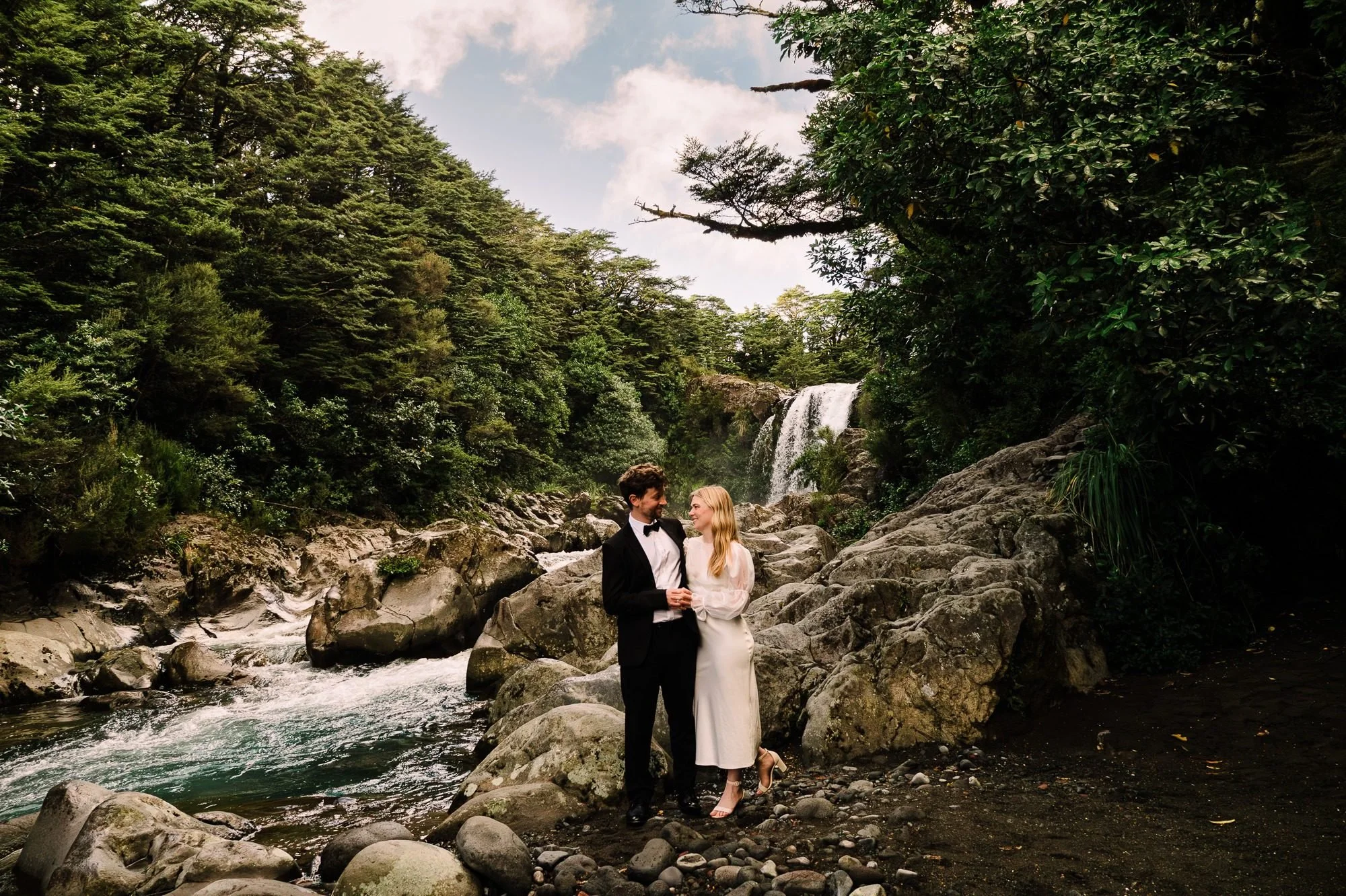 Styled elopement couple standing by the river below Tawhai Falls during a New Zealand elopement photography session by Kevin M Films.