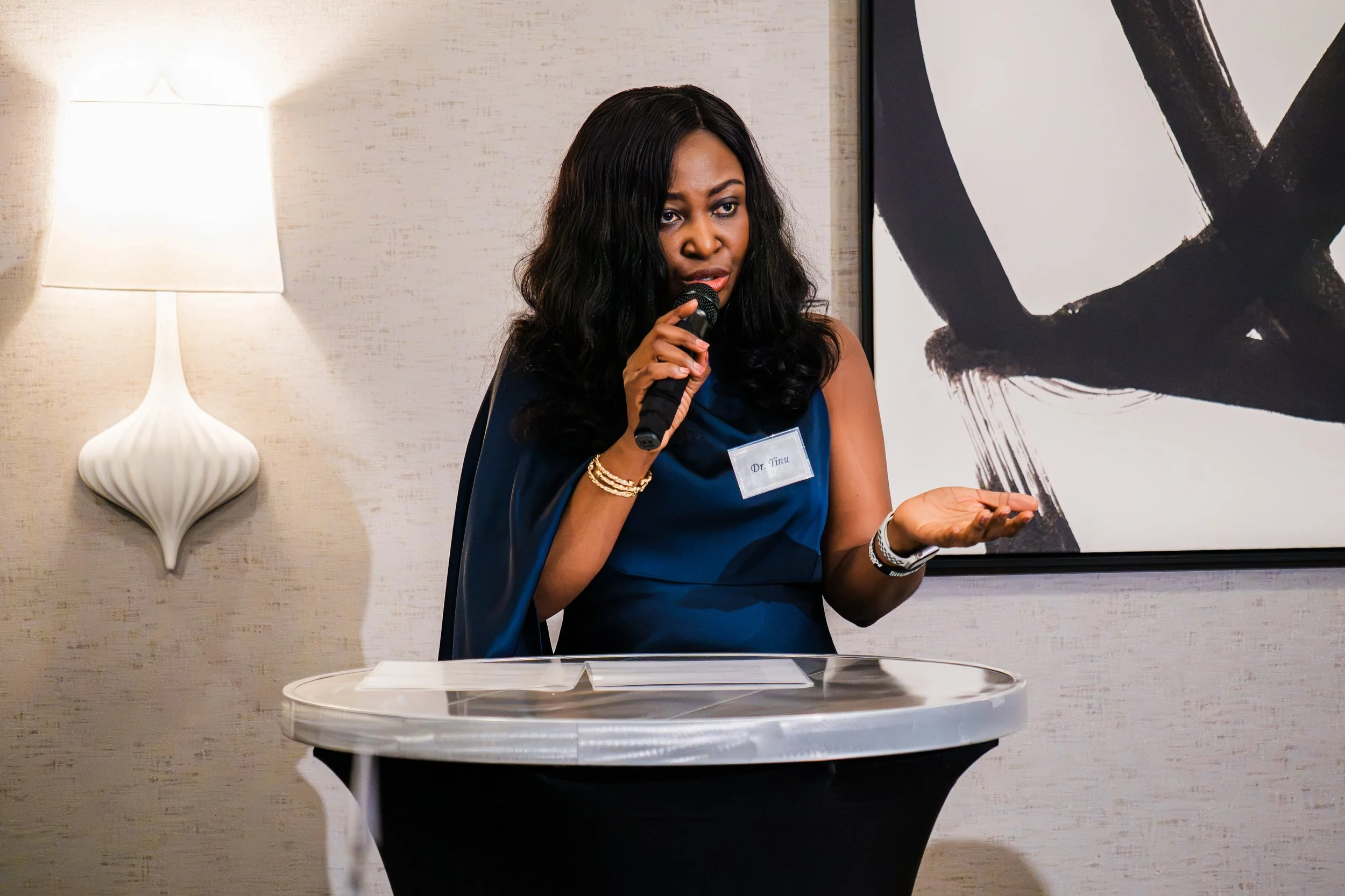 Speaker in a blue dress addressing guests with a handheld microphone at the Synergy Family Medical Clinic dinner.