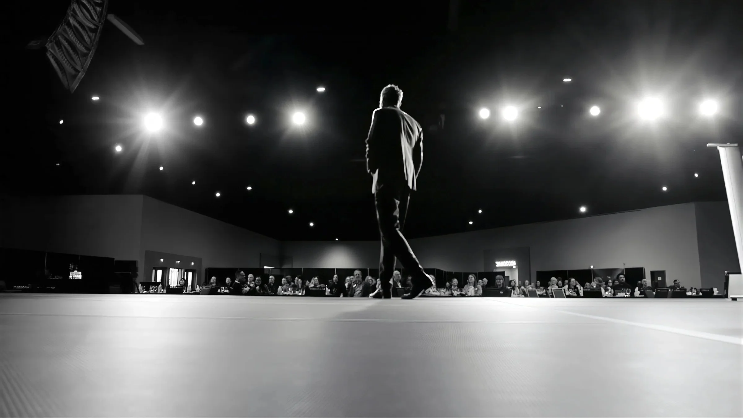 Black-and-white photo of a speaker onstage facing a seated audience beneath bright event lights.