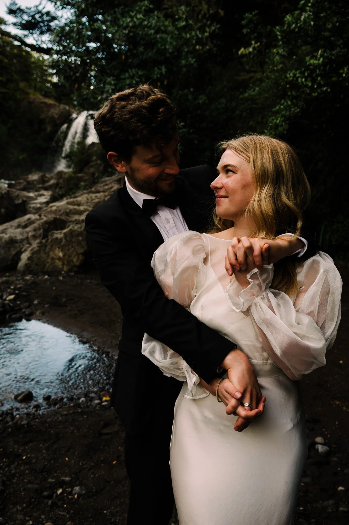 Styled elopement couple embracing beside the river at Tawhai Falls during a New Zealand elopement photography session by Kevin M Films.