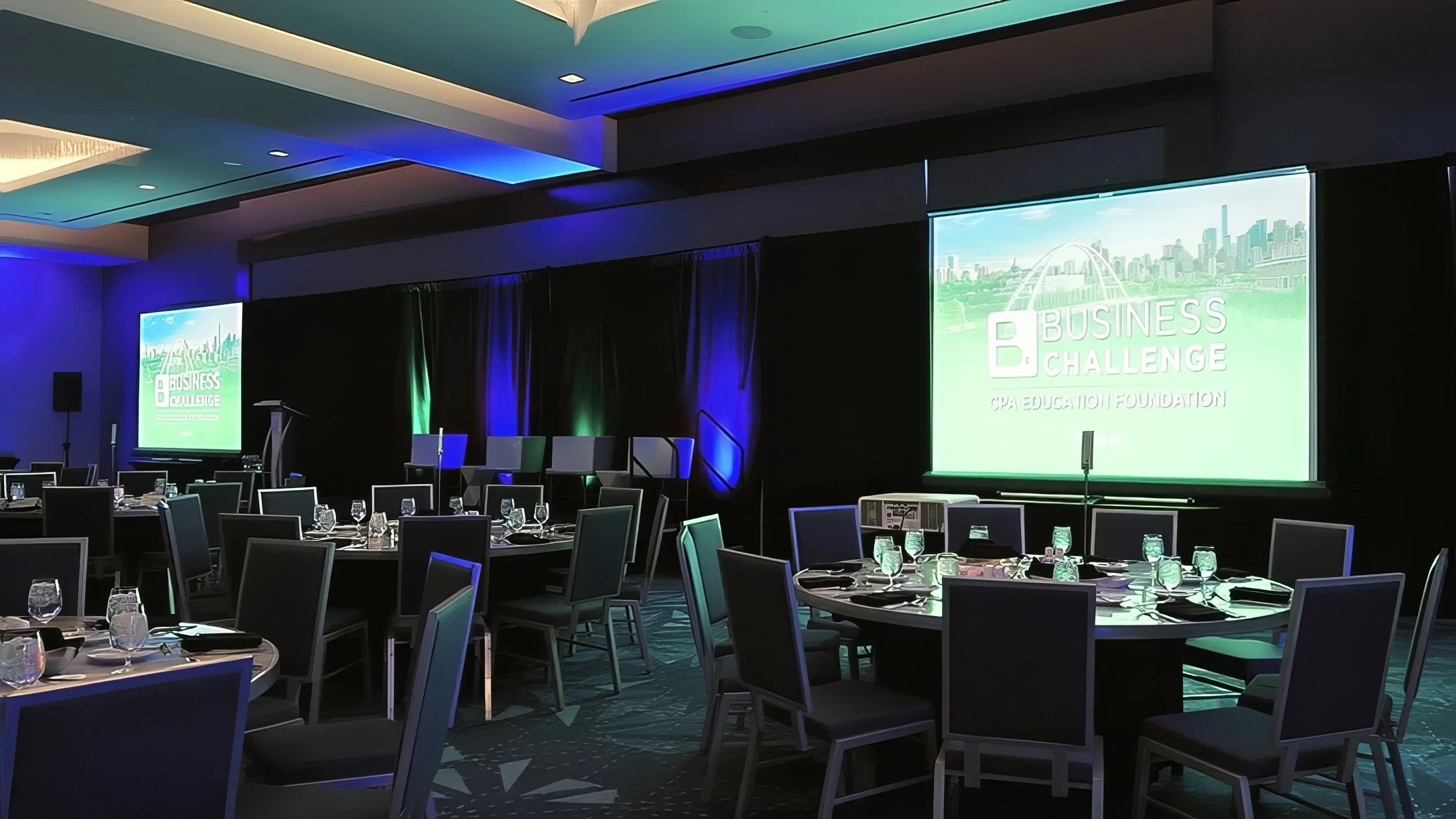 Banquet room set for the CPA Education Foundation Business Challenge, with round dining tables, a stage, projection screens, and blue and green lighting.