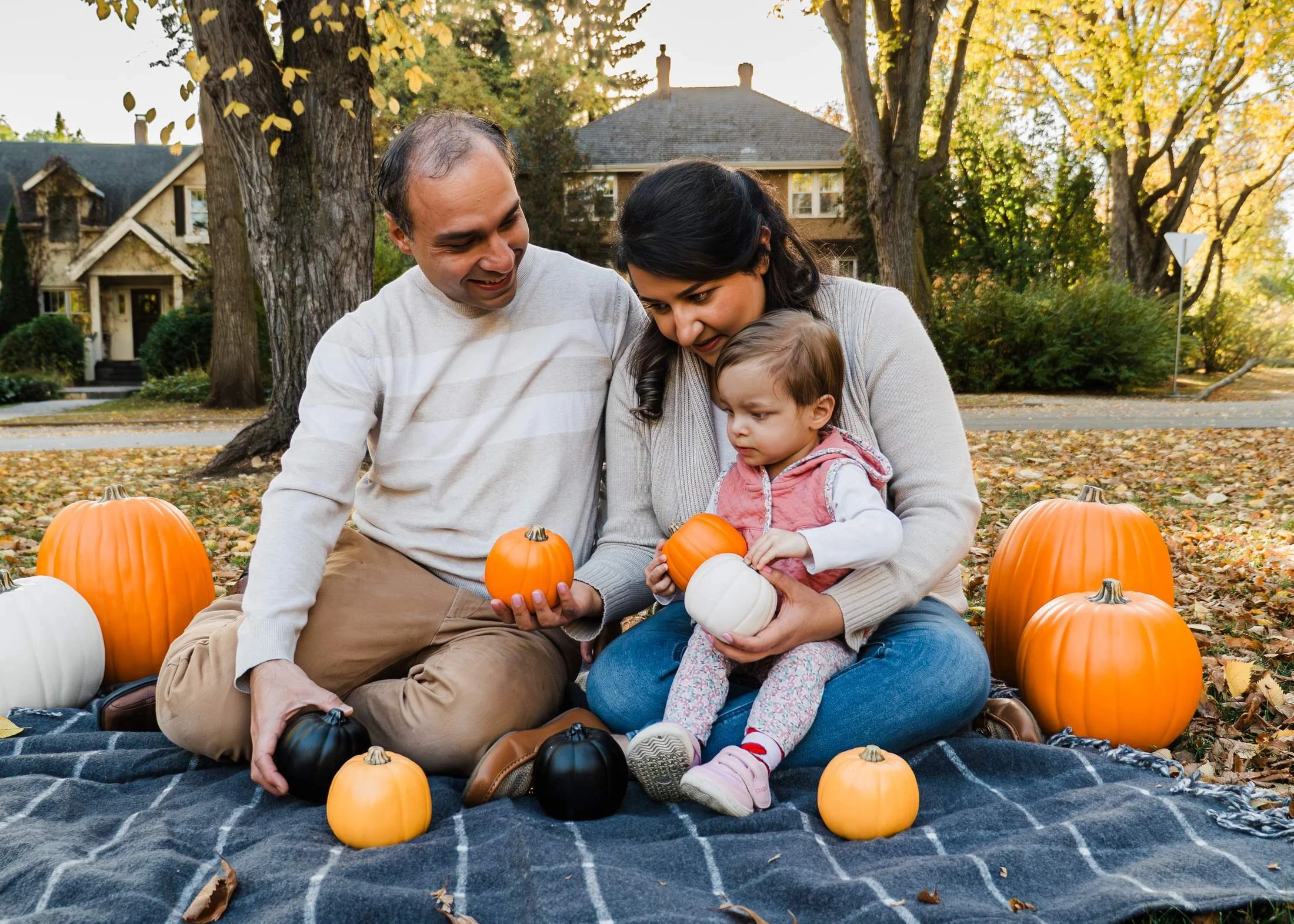 Parents sitting with their young daughter on a plaid blanket, holding small pumpkins beneath golden fall trees.