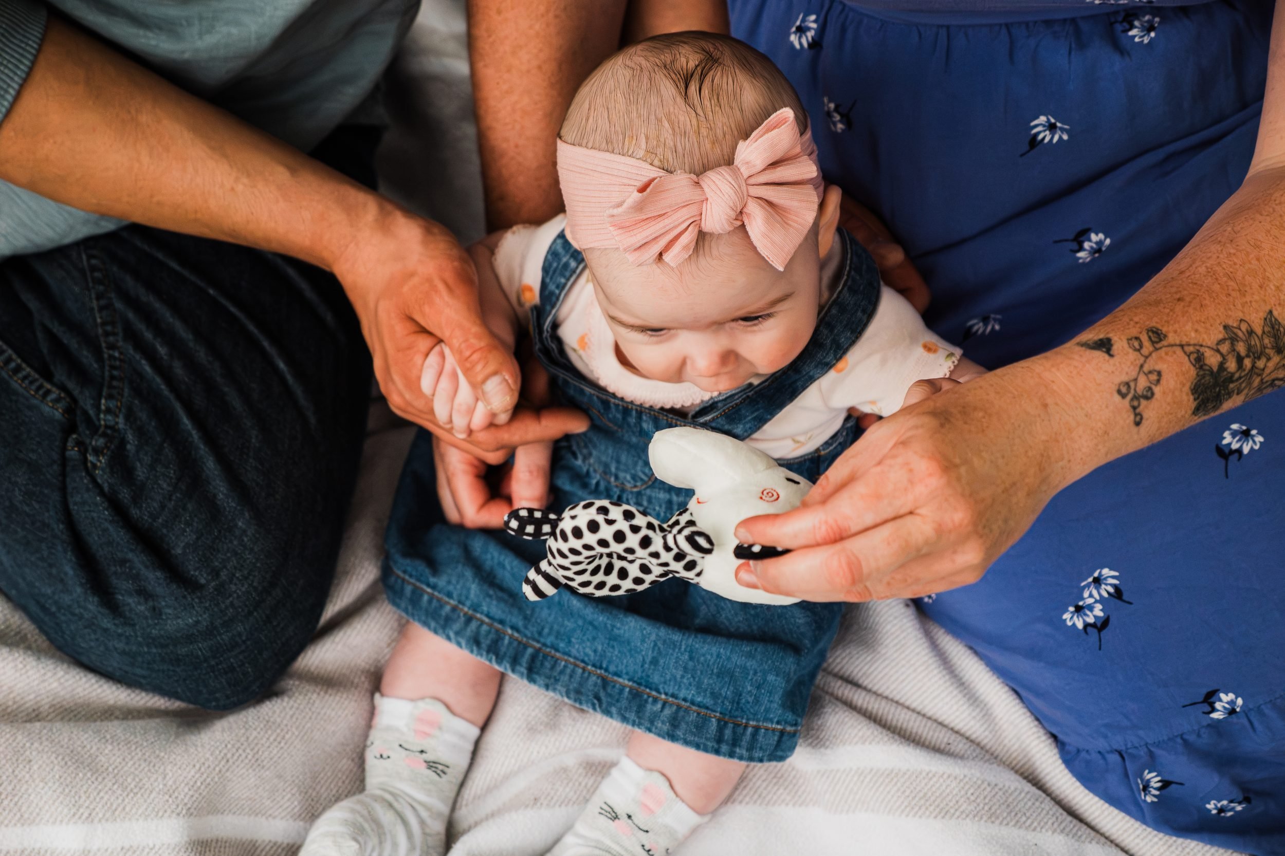 Baby wearing a pink bow and denim dress, sitting between their parents as they hold the baby’s hand and offer a soft toy