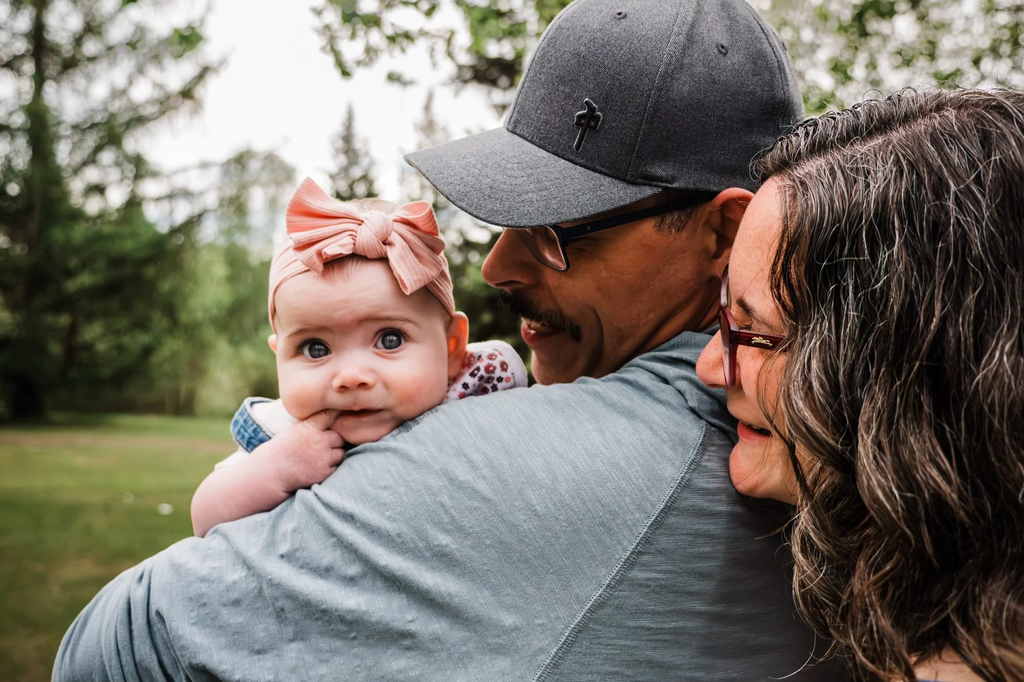 Outdoor family portrait of parents cuddling their baby, who wears a pink bow and looks toward the camera.