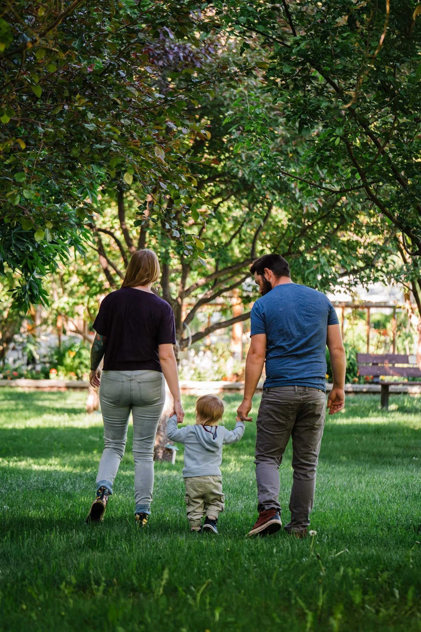Parents holding their young child’s hands as they walk across the grass beneath leafy trees in a sunlit garden.