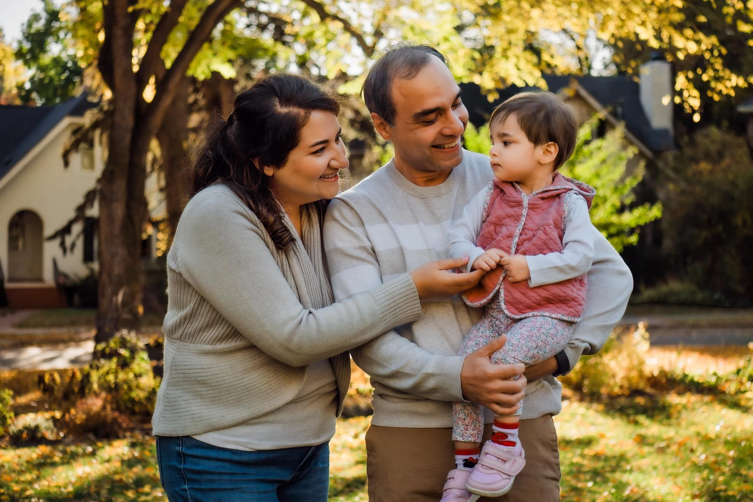 Parents smiling at their young daughter beneath golden fall trees.