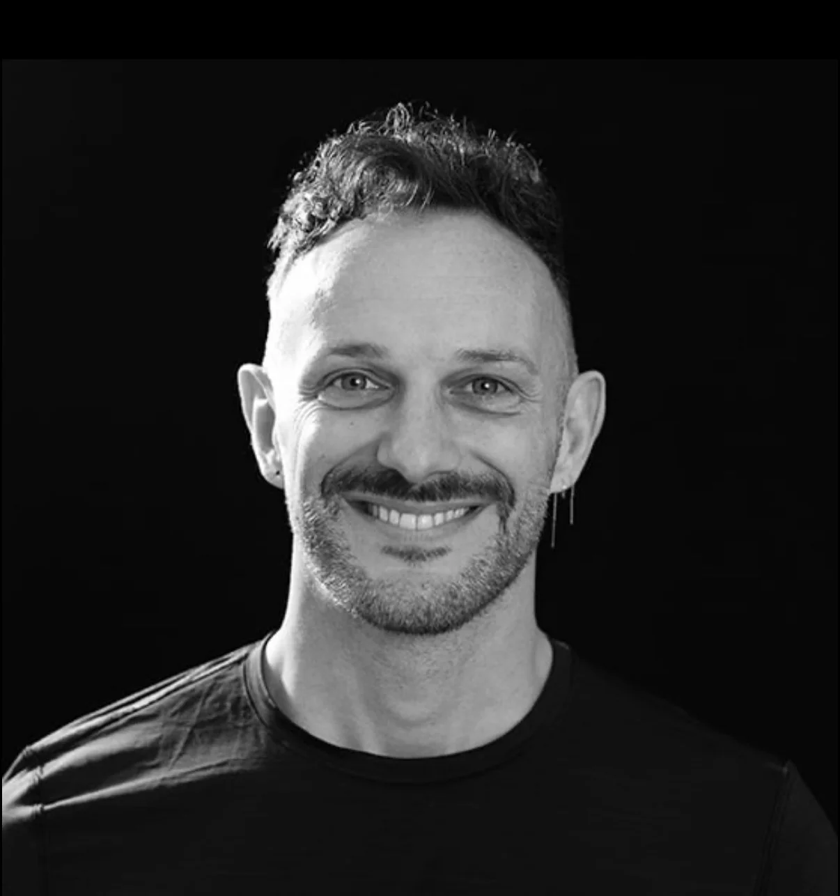 Black and white portrait of a Justin with short curly hair, a beard, and an earring, wearing a dark shirt, against a black background.