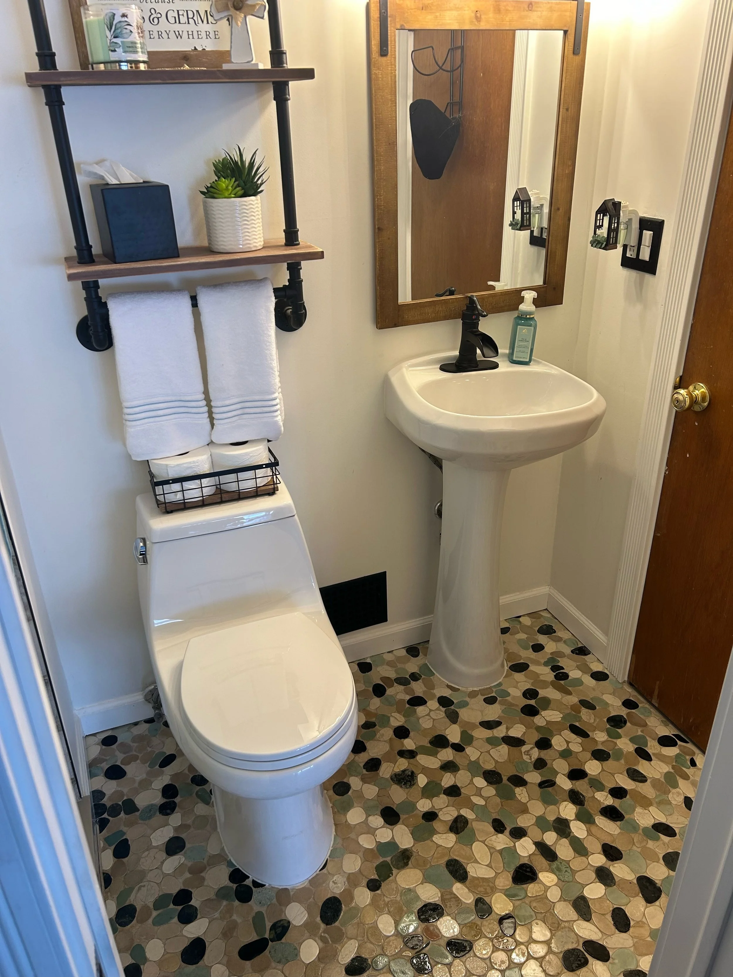 Small bathroom with a white toilet, a pedestal sink with a black faucet, a small mirror with a wooden frame, and a wooden door. Above the toilet is a black shelf with two levels holding a potted plant, a tissue box, and decorative items. On the wall to the right of the sink are two small black wall-mounted shelves with plants. The floor has a multicolored pebble tile pattern.