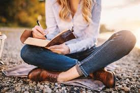 Person sitting cross-legged outdoors, writing in a notebook on a blanket.