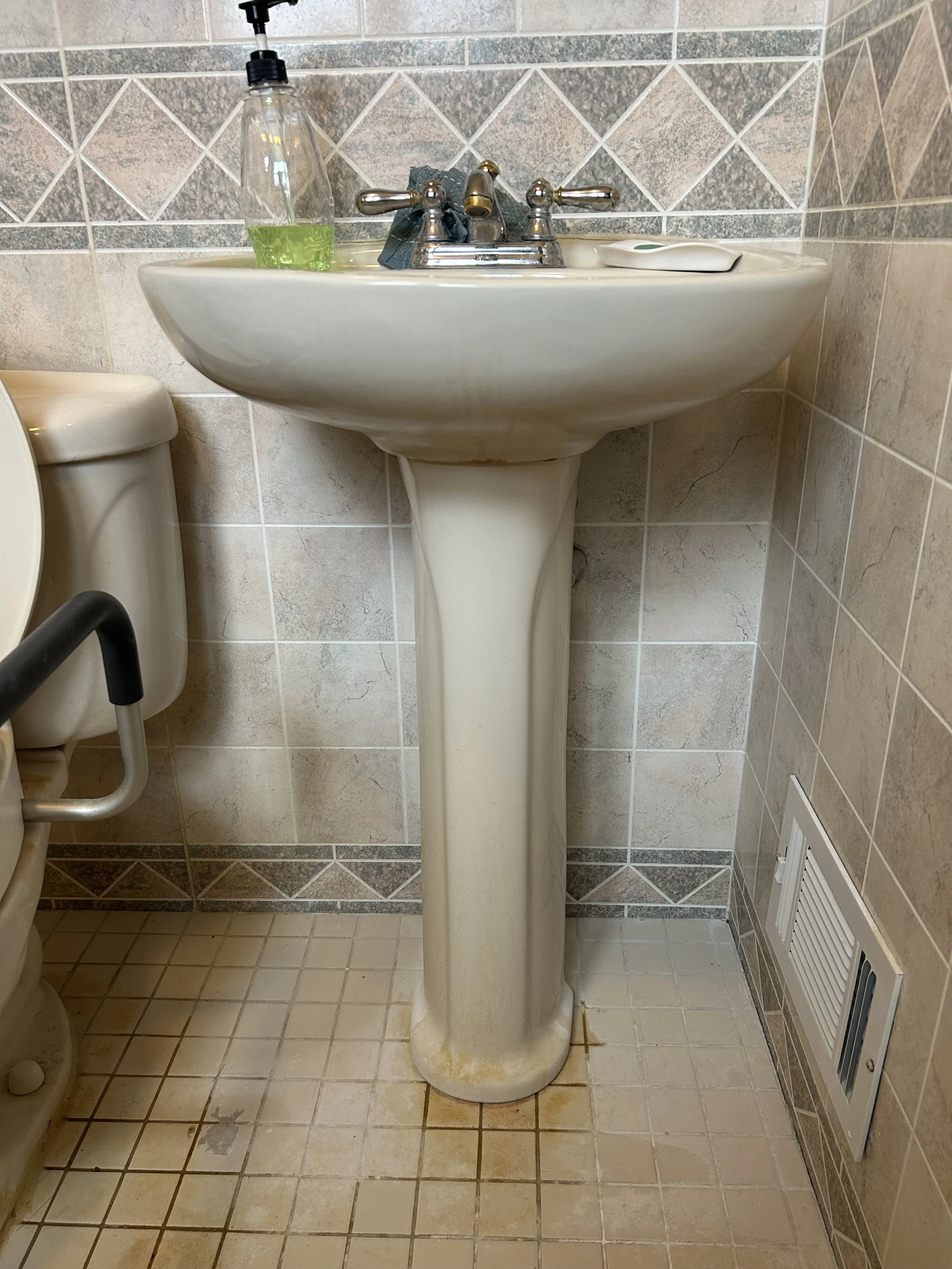 Bathroom sink with soap, hand sanitizer, and soap dish, with a toilet in the background and tiled walls and floor.