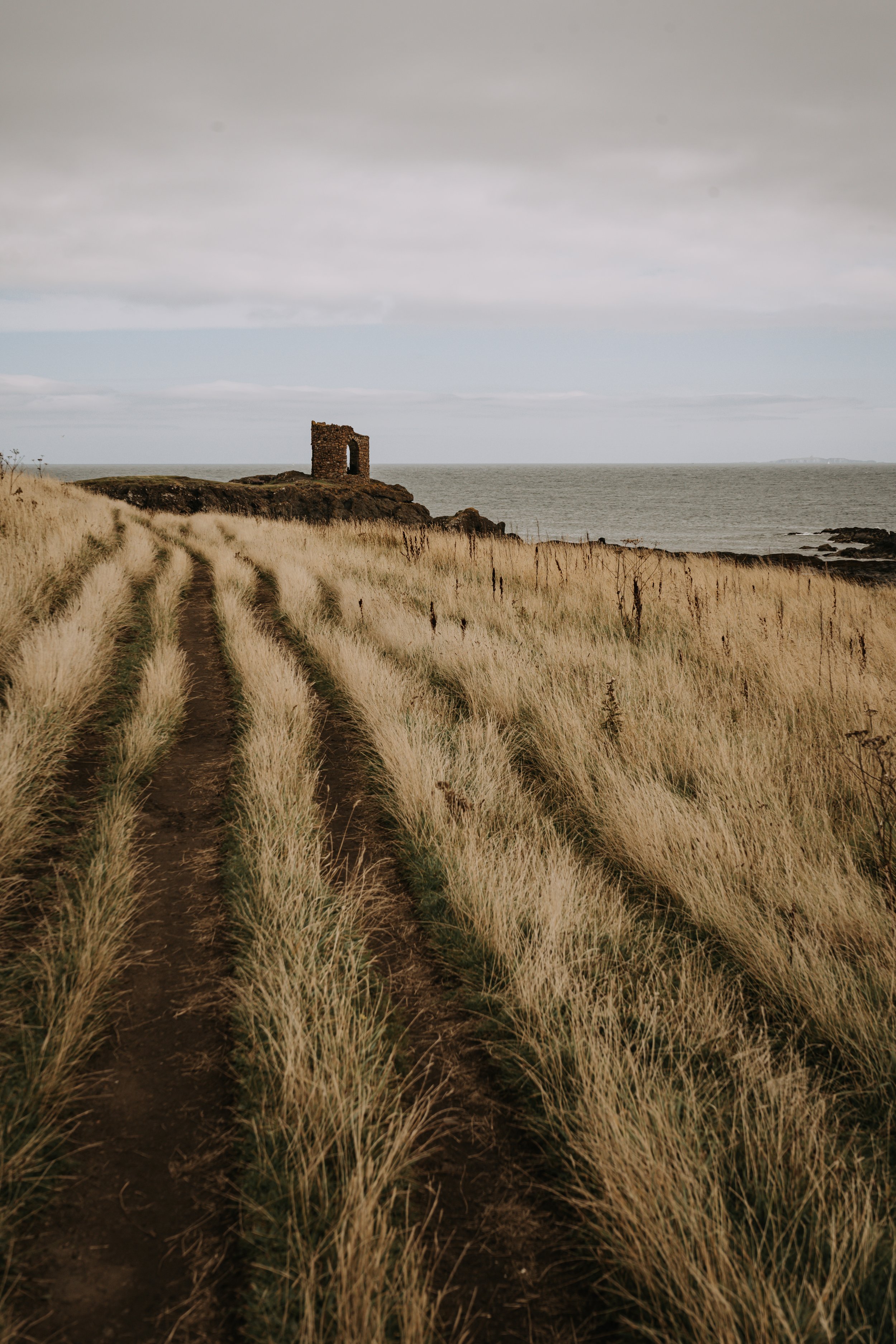 Coastal path leading to a stone ruin overlooking the ocean, surrounded by tall grass under an overcast sky.