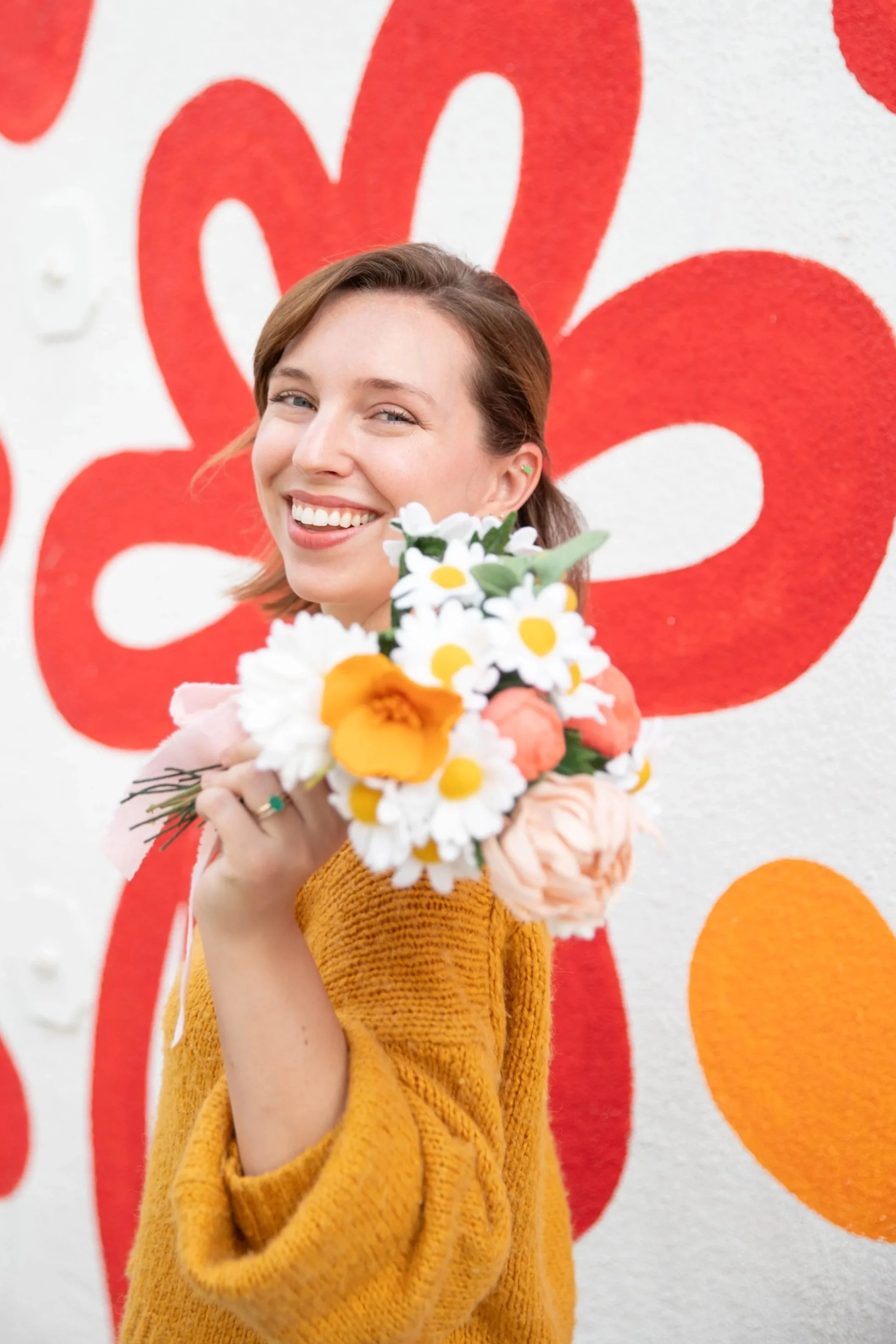 A woman in a mustard yellow sweater holding a bouquet of daisies and other flowers, smiling and standing in front of a colorful wall with red and orange abstract shapes.