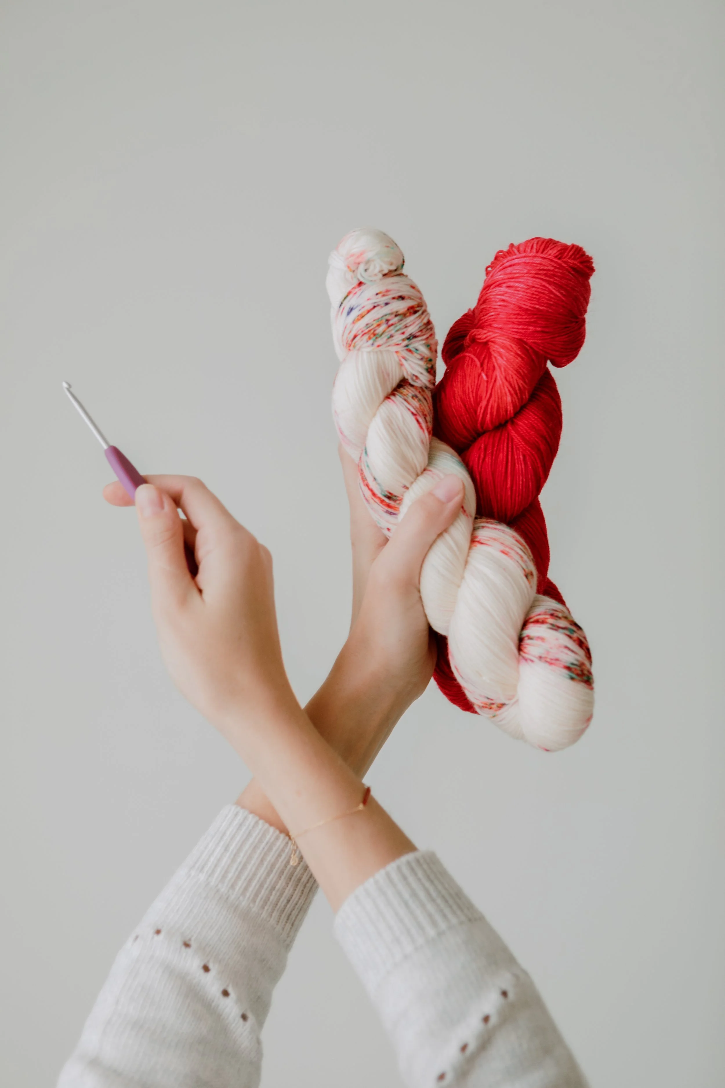 Person holding two skeins of yarn, one red and one multicolored, with a crochet hook in hand, against a plain light background.