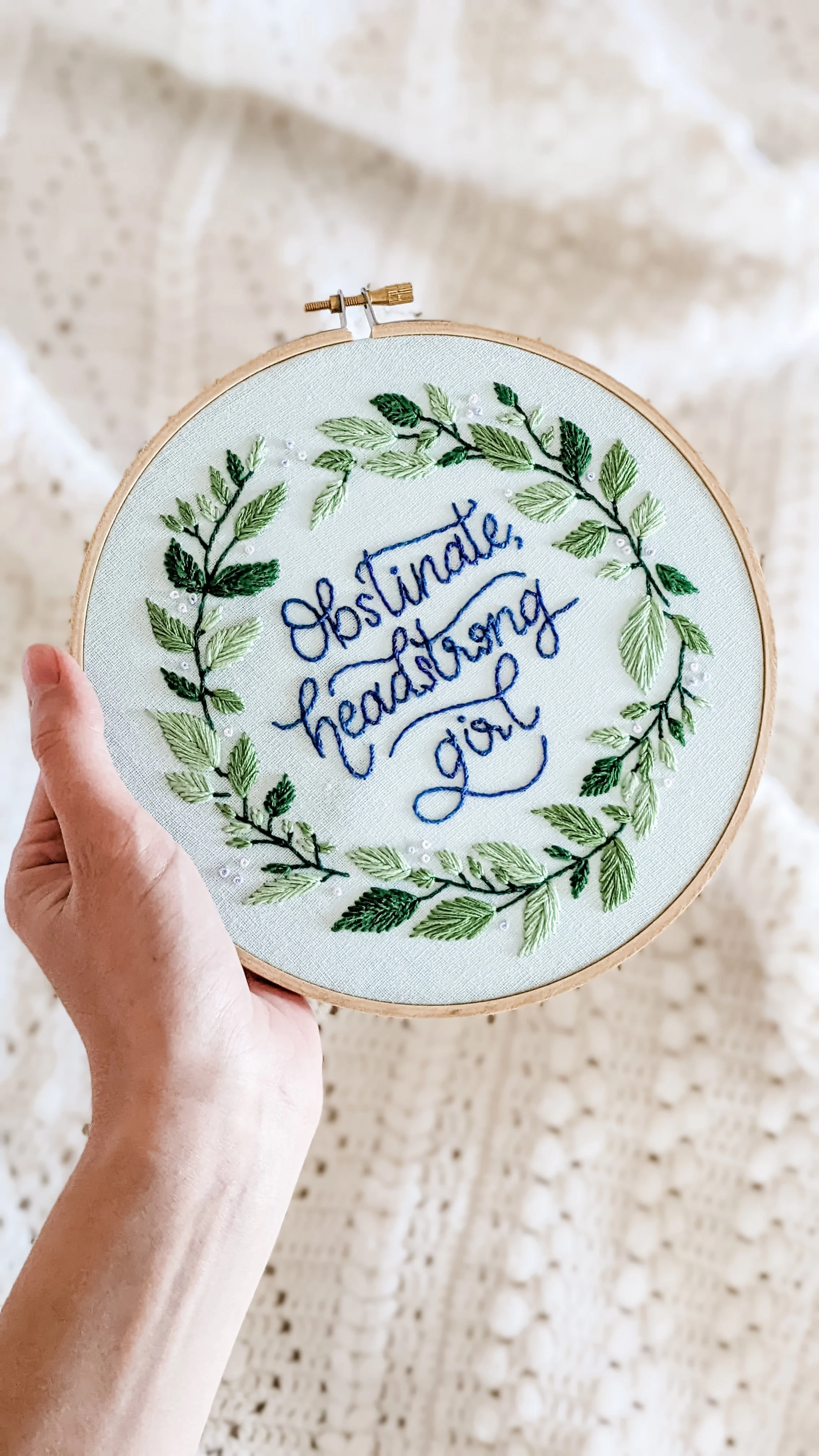 Embroidery hoop with a stitched quote 'Obstinate, headstrong girl' surrounded by green embroidered leaves.