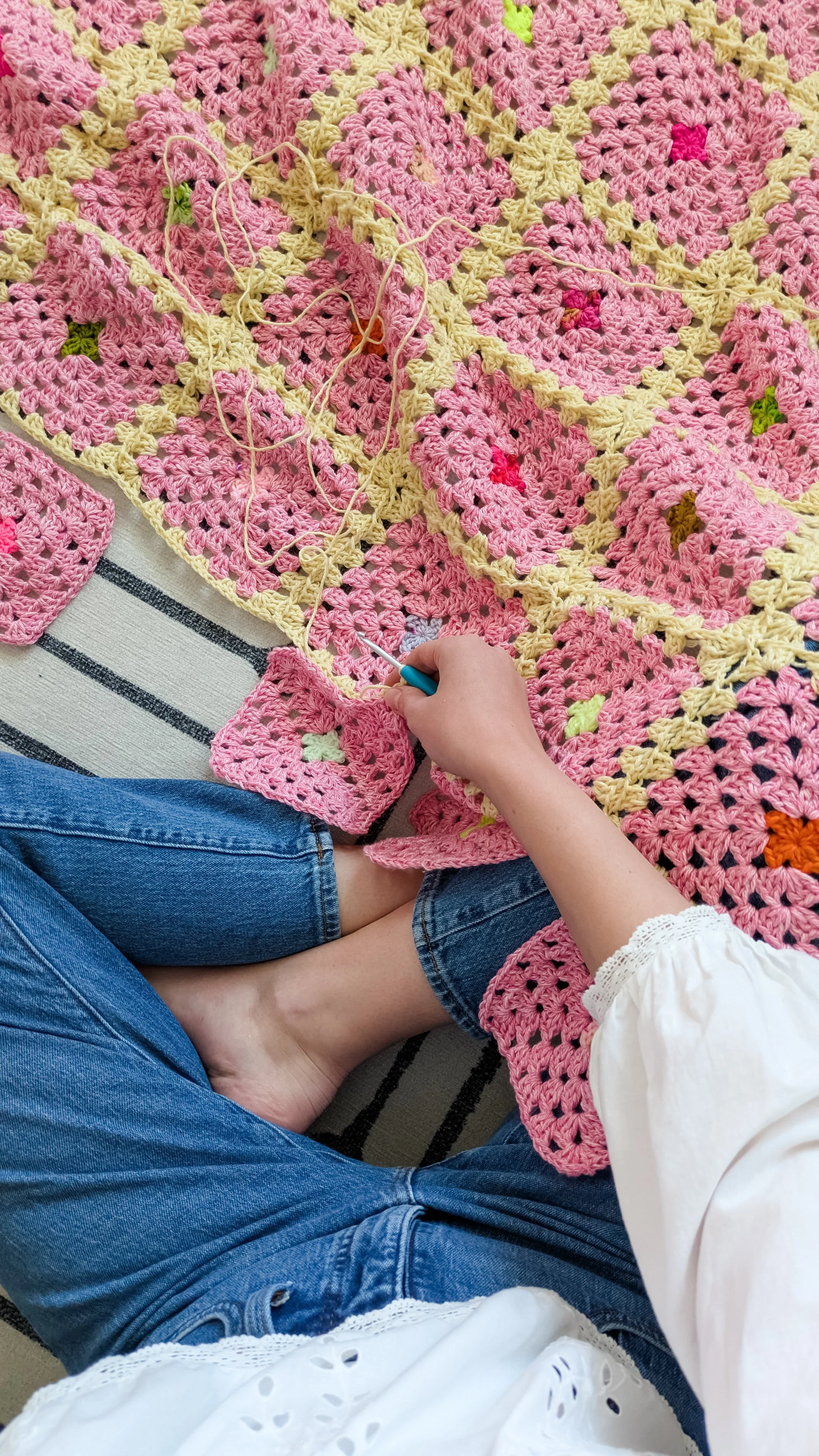 Person crocheting a pink and yellow blanket on the floor, wearing blue jeans and a white blouse with eyelet details.