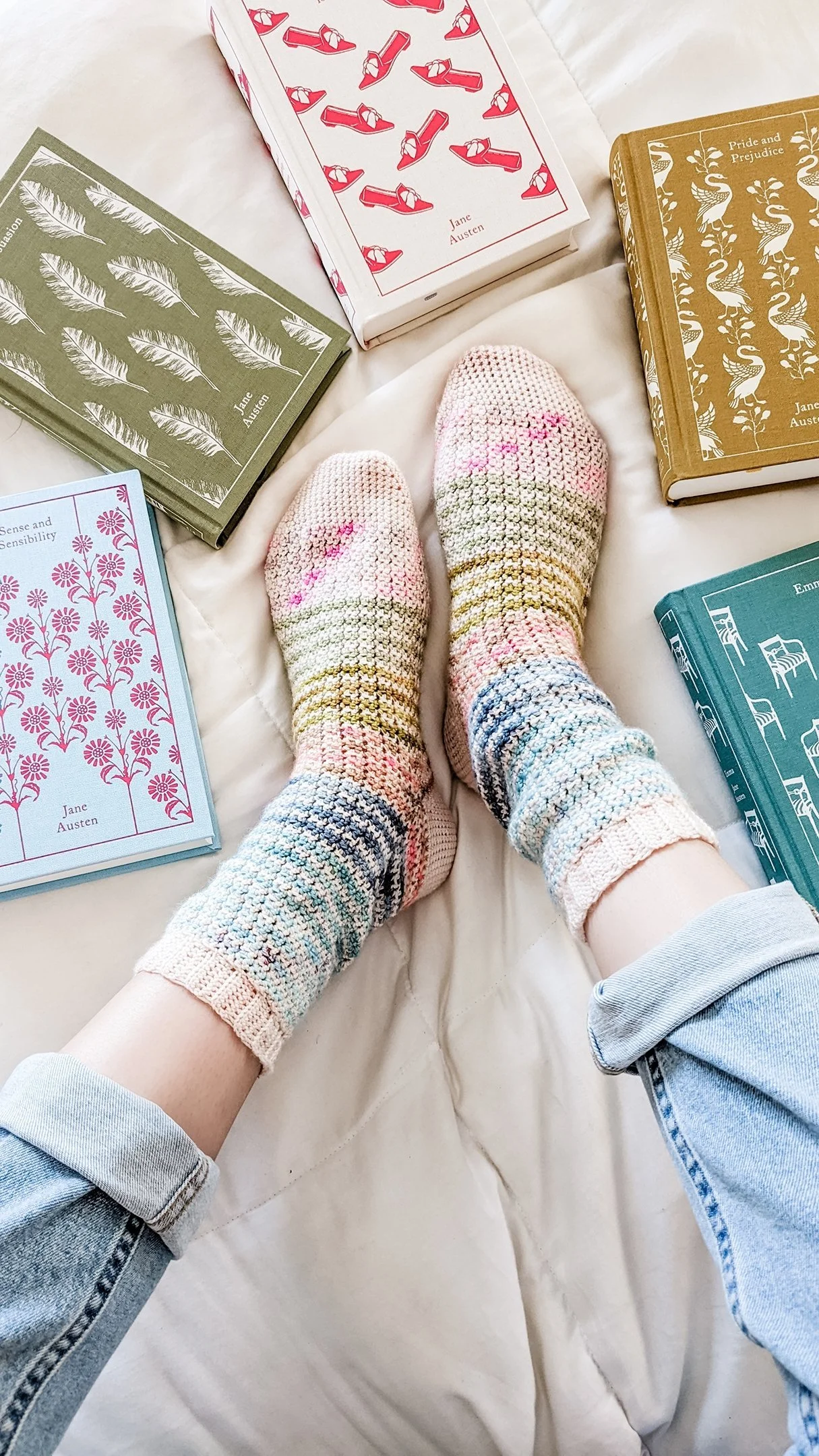 Person wearing multicolored crocheted socks with rolled-up light blue jeans, lying on a beige bed surrounded by several colored books with Jane Austen titles.