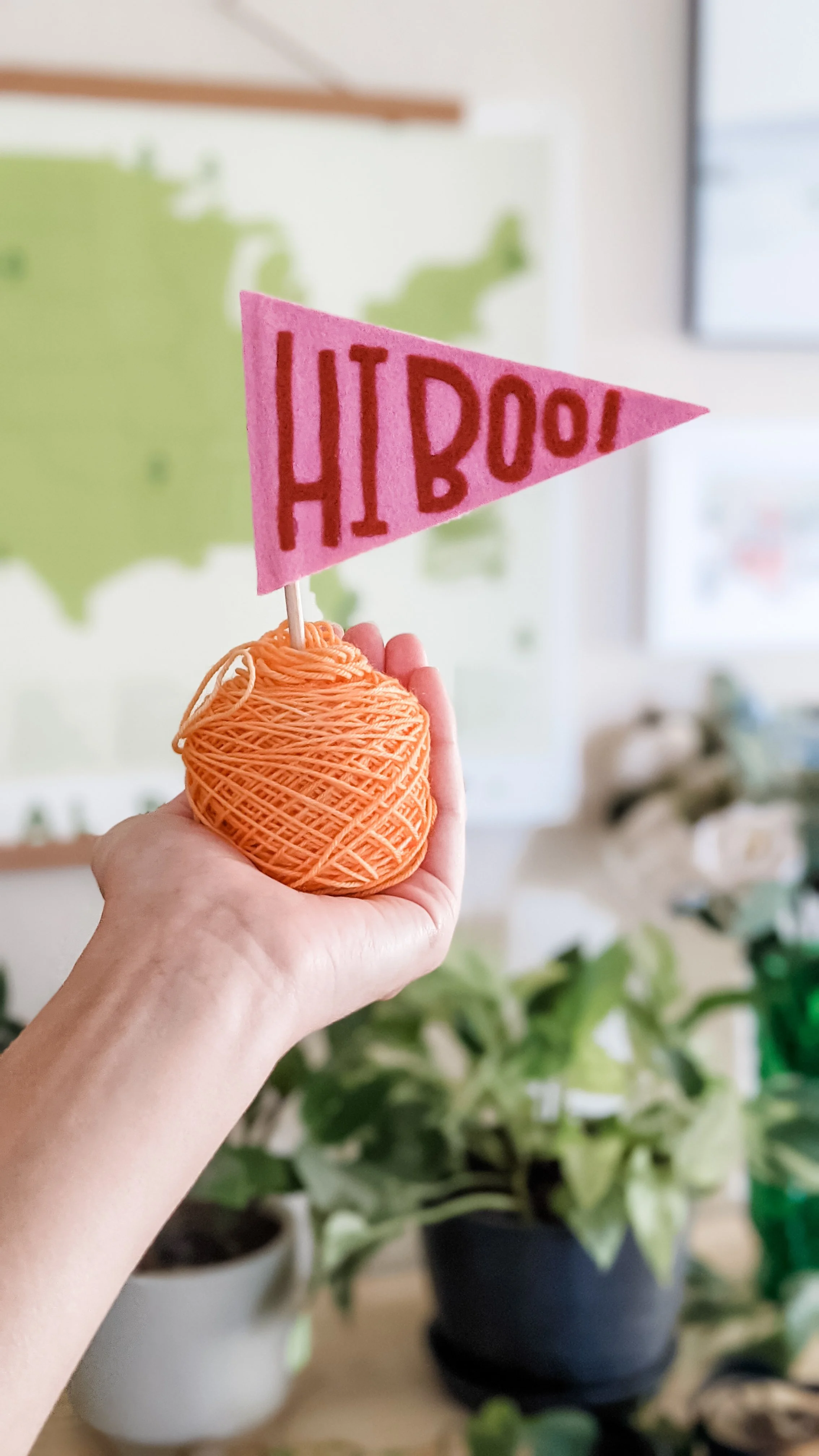 A hand holding a small ball of orange yarn with a pink felt flag on top that has red letters spelling 'HI BOO!'. In the background, there are potted plants and a blurred map of the United States on the wall.