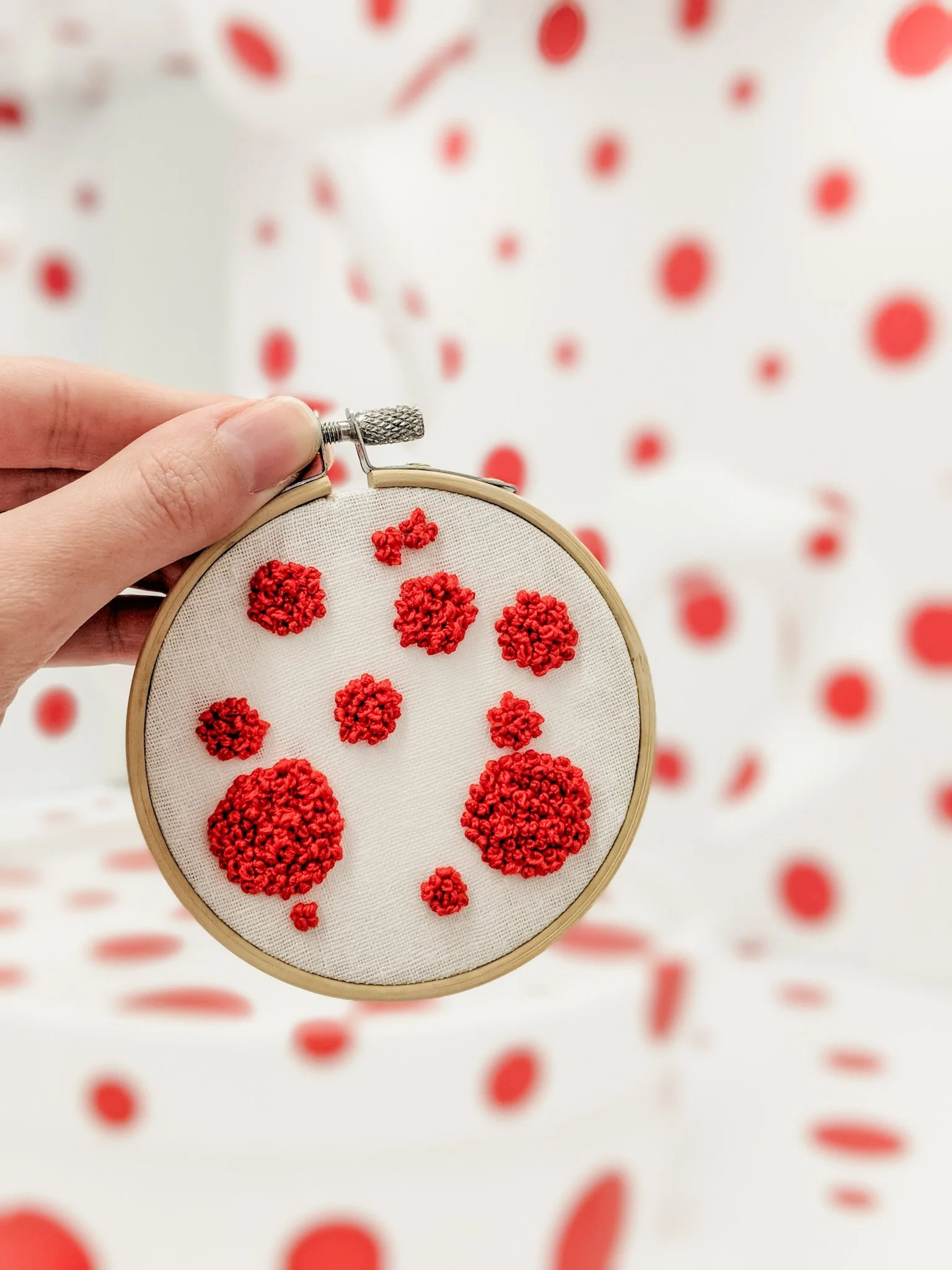 Close-up of embroidery hoop with red dot patterns on white fabric, held by a person's hand against a blurred background of similar red patterns.