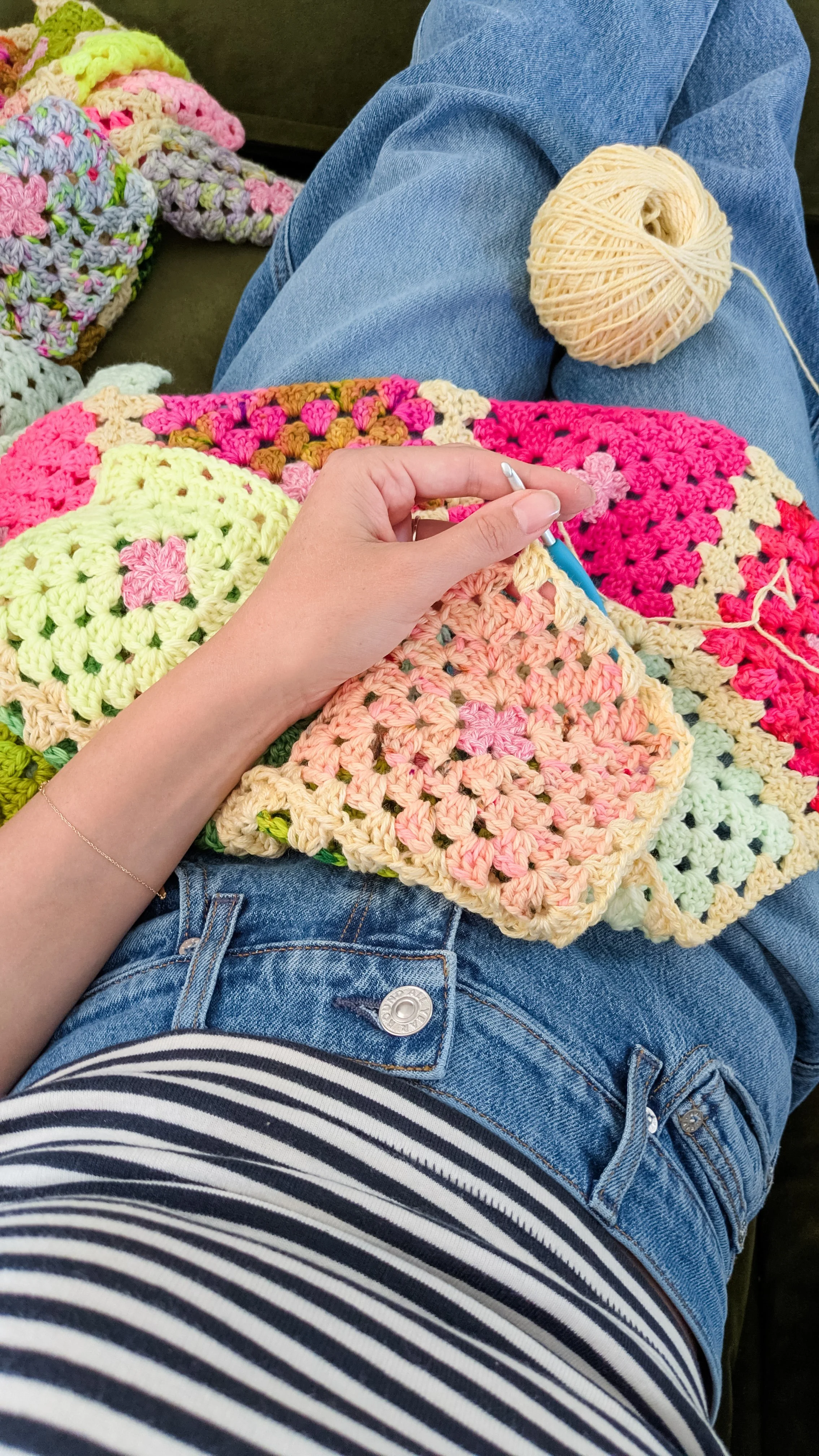 A person sitting with their legs crossed, surrounded by colorful crochet blankets. They are holding a crochet hook, working on a crochet project. There is a ball of yellow yarn resting on their lap and they are wearing blue jeans and a striped shirt.