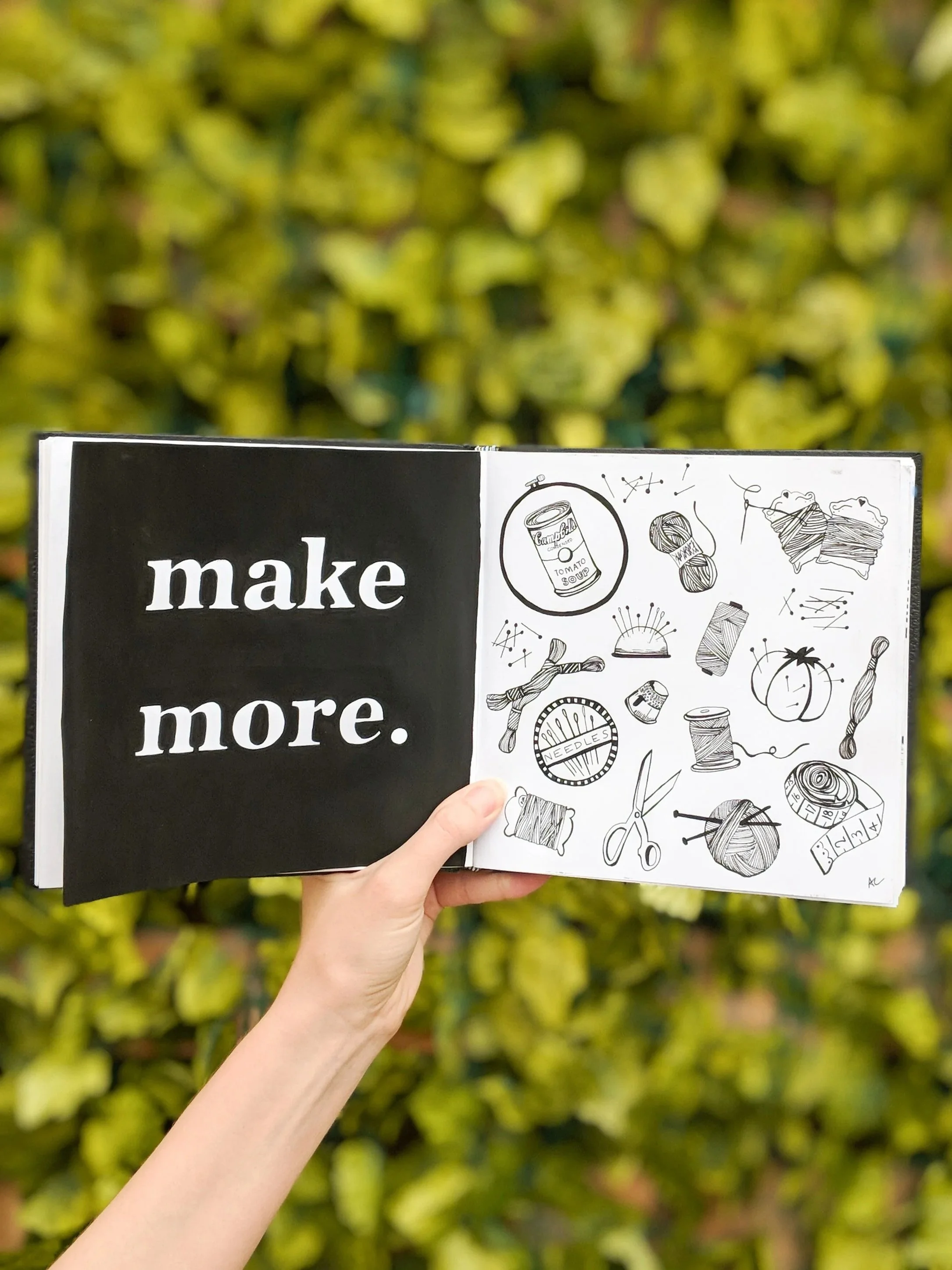 A person holding an open black and white illustrated book with the words "make more" on the left page and various doodles related to knitting, sewing, and crafting on the right page. The background features blurred green foliage.