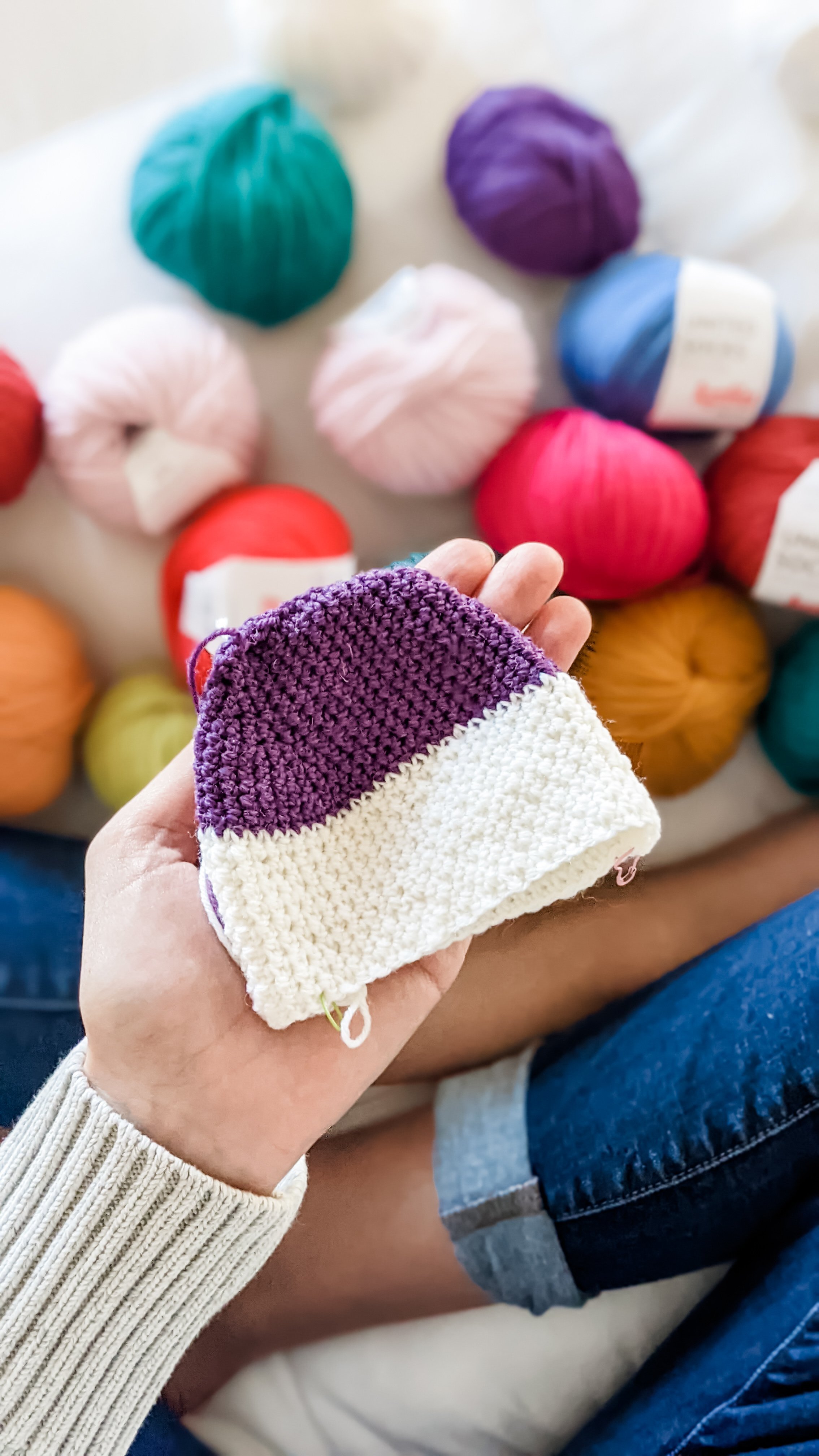 A person holding a small, two-toned crochet sock item, with multicolored yarn balls spread out in the background.