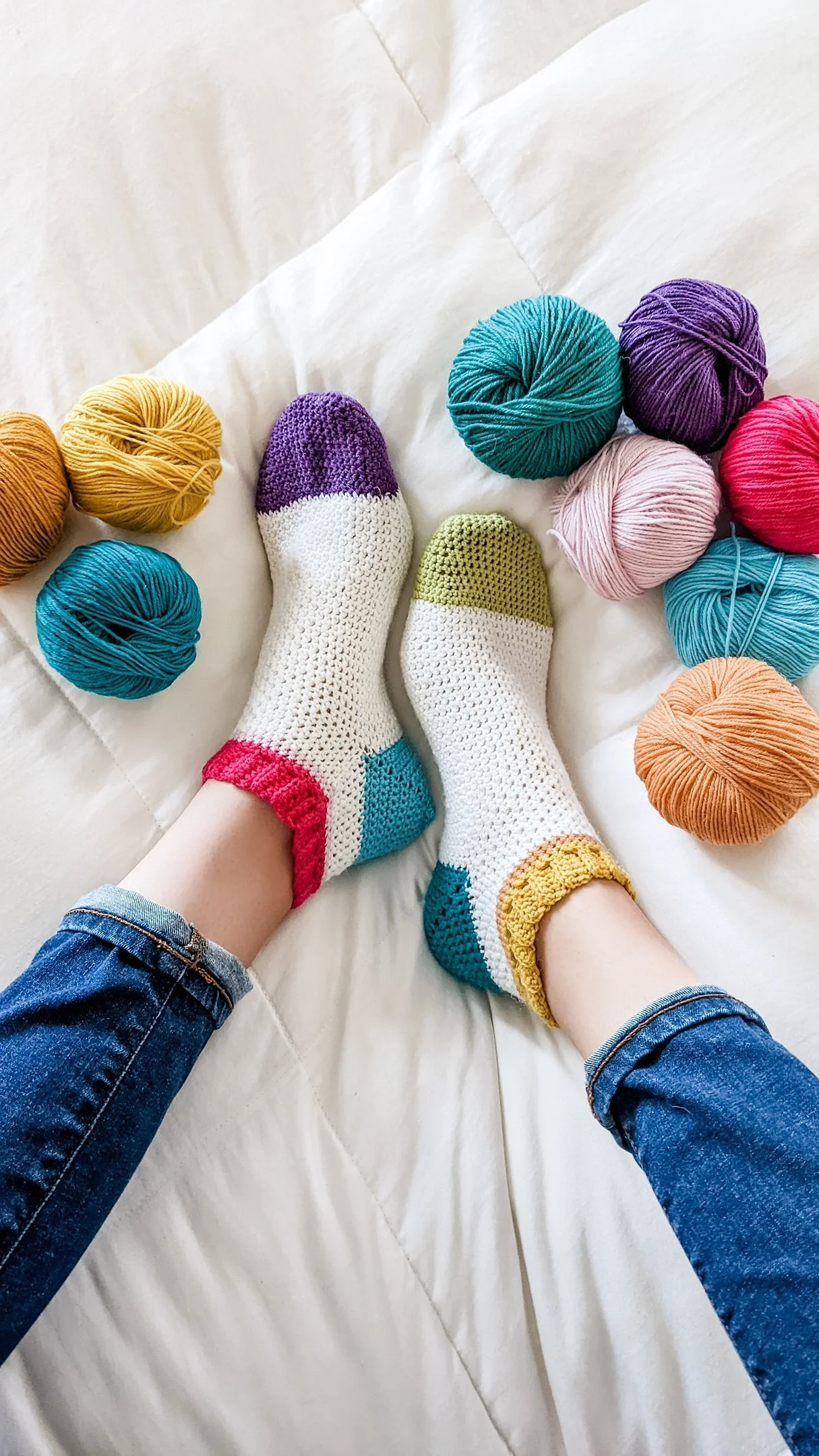 Person wearing colorful crochet socks and blue jeans, lying on a white bed surrounded by balls of colorful yarn in yellow, purple, turquoise, pink, and orange.