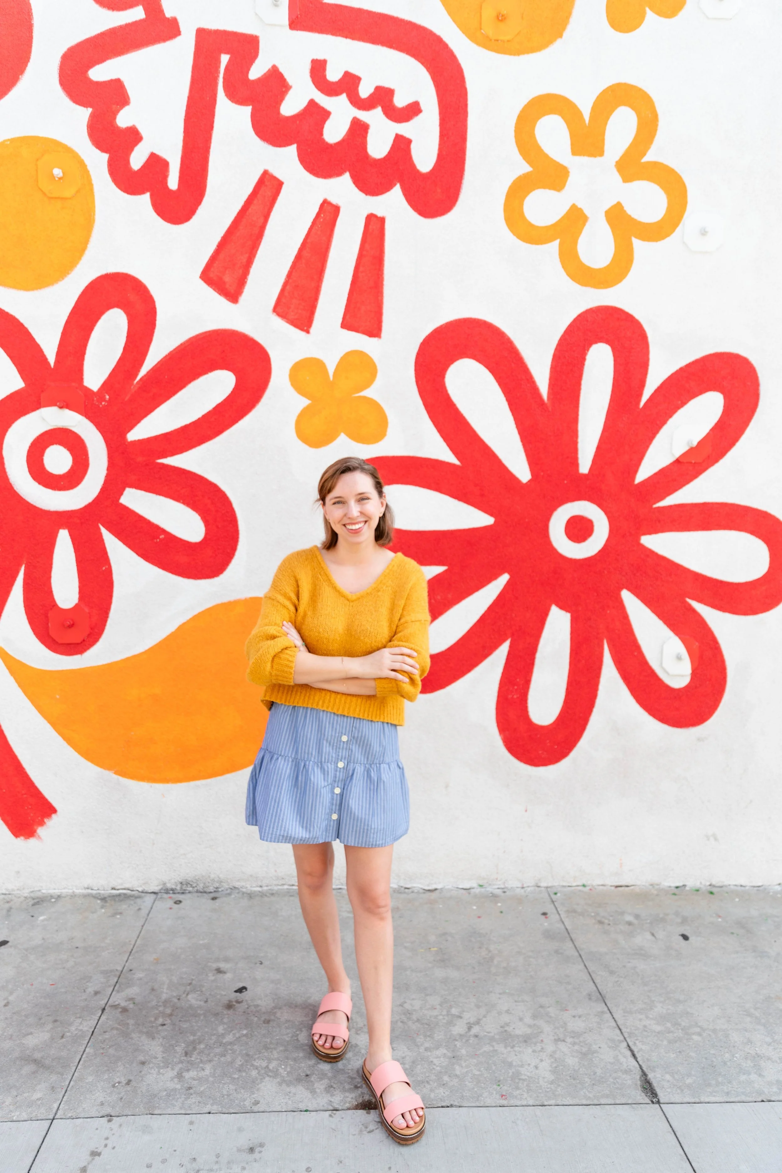 A young woman stands in front of a vibrant mural featuring large, colorful flowers in red and orange on a white wall. She is smiling, with her arms crossed, wearing a mustard yellow sweater, a blue and white striped skirt, and pink platform sandals.