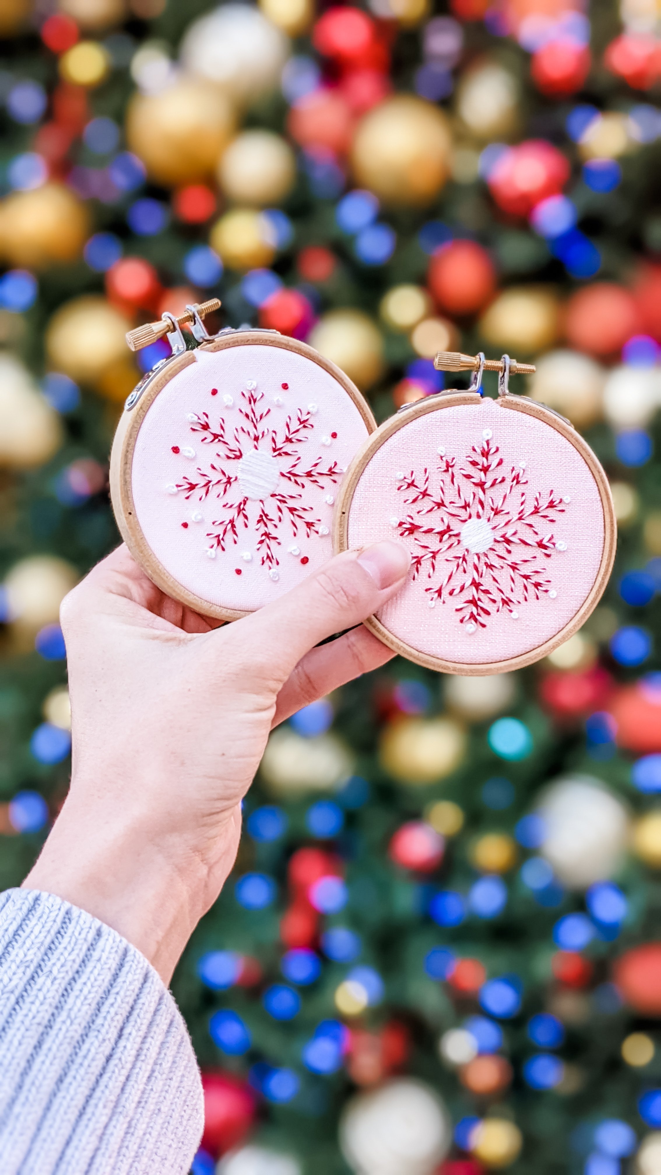 Hand holding two pink fabric embroidery hoop ornaments with red and white snowflake design, in front of a decorated Christmas tree with gold, red, and purple ornaments and colorful lights.