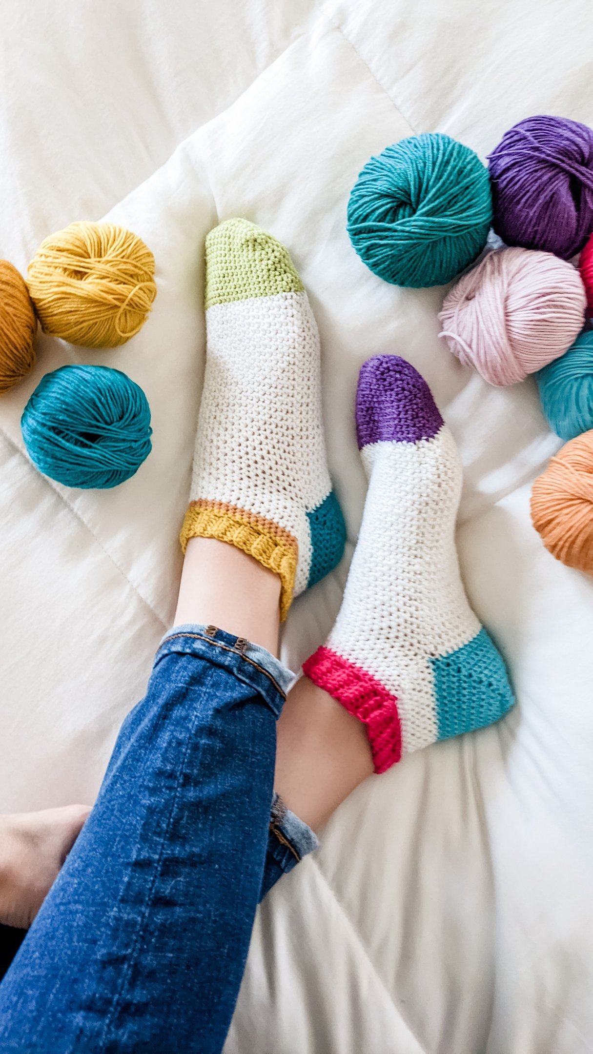 Person wearing colorful crocheted socks and blue jeans, lying on a white bed surrounded by balls of yarn in various colors.