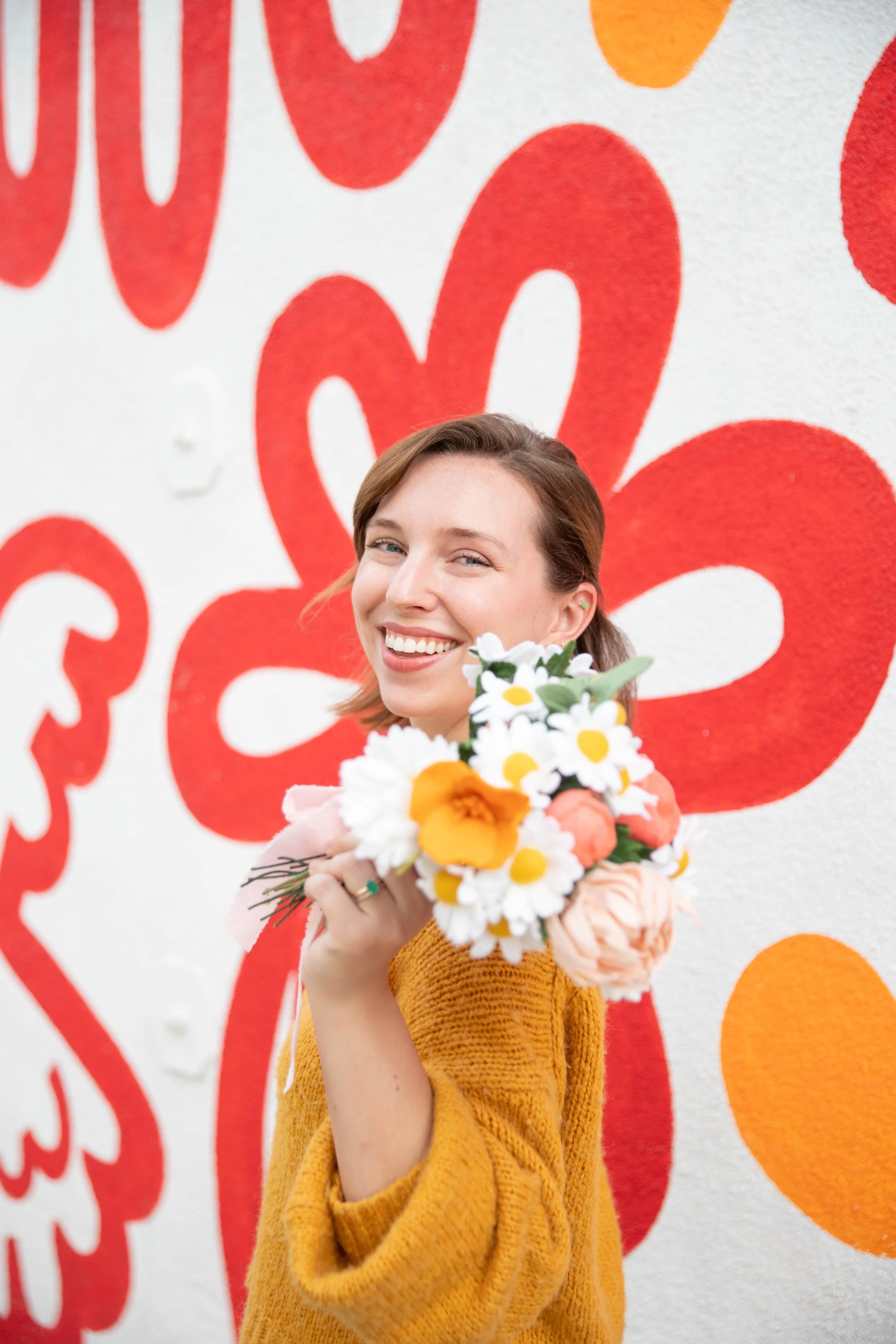 A smiling woman with short brown hair holding a bouquet of white and yellow flowers, wearing a mustard-colored sweater, standing in front of a colorful mural with red and orange flower shapes.