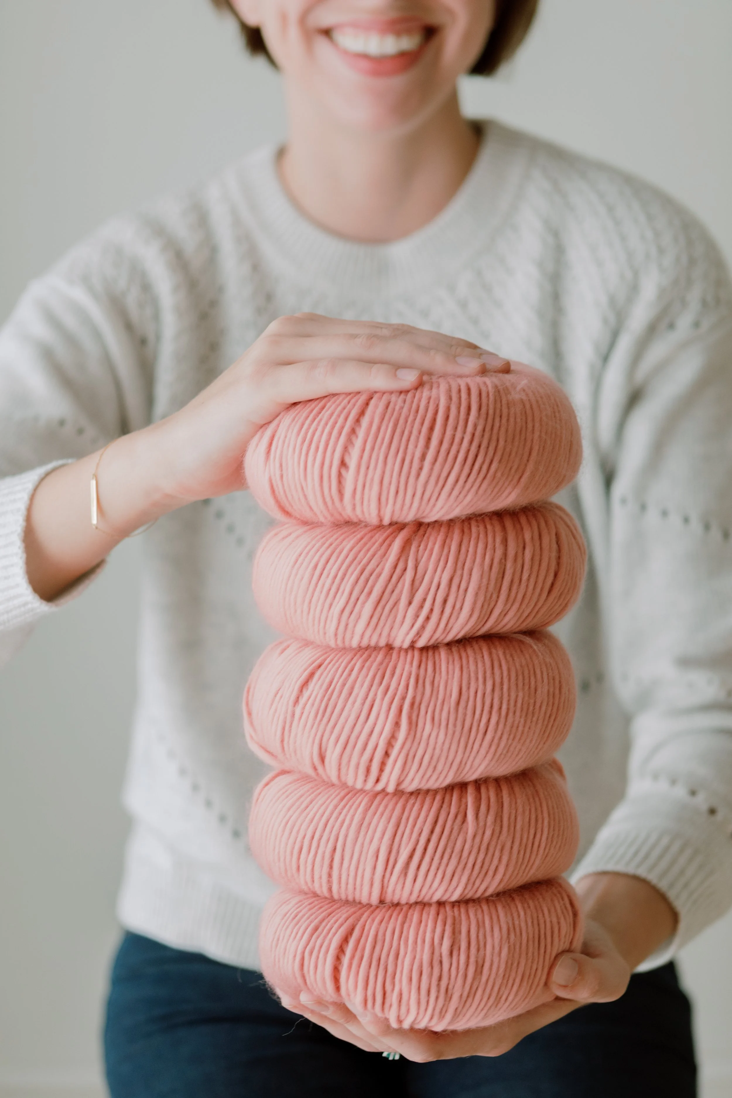 A person smiling while holding a stack of pink, ridged, velvet yarn skeins.