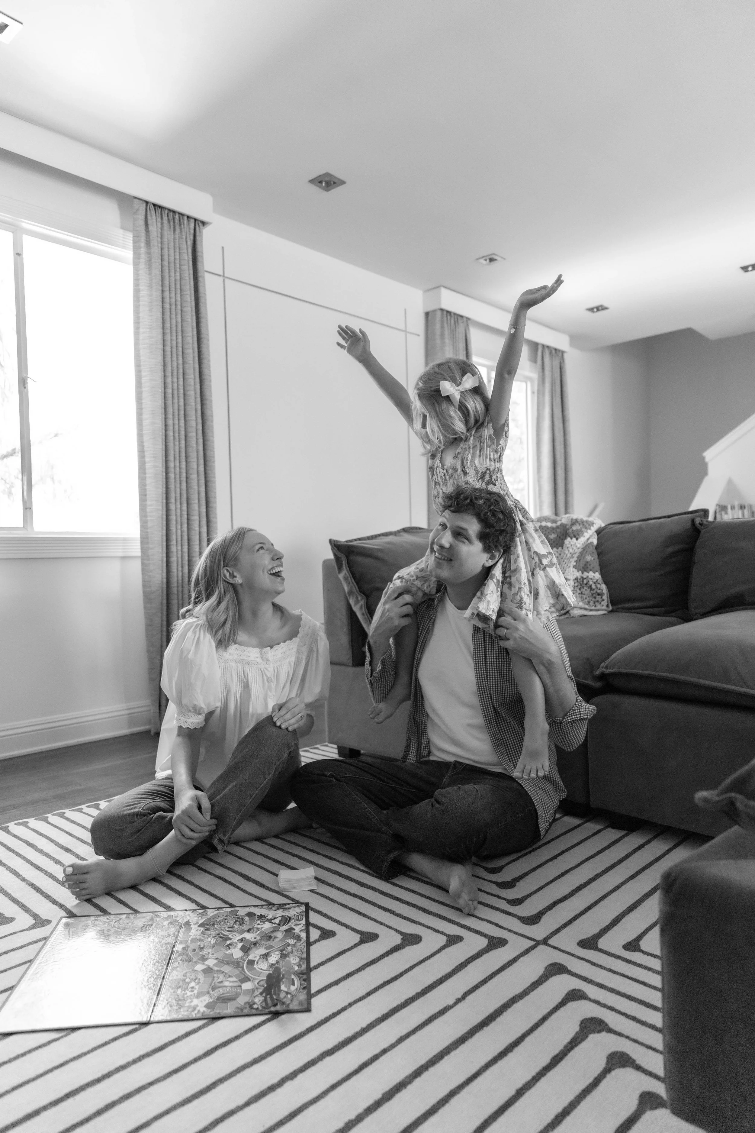 A family, with a father, mother, and  young daughter, playing together on a striped area rug in a living room. The father is sitting cross-legged with one daughter seated on his shoulders. The mother is kneeling in front of them, smiling.