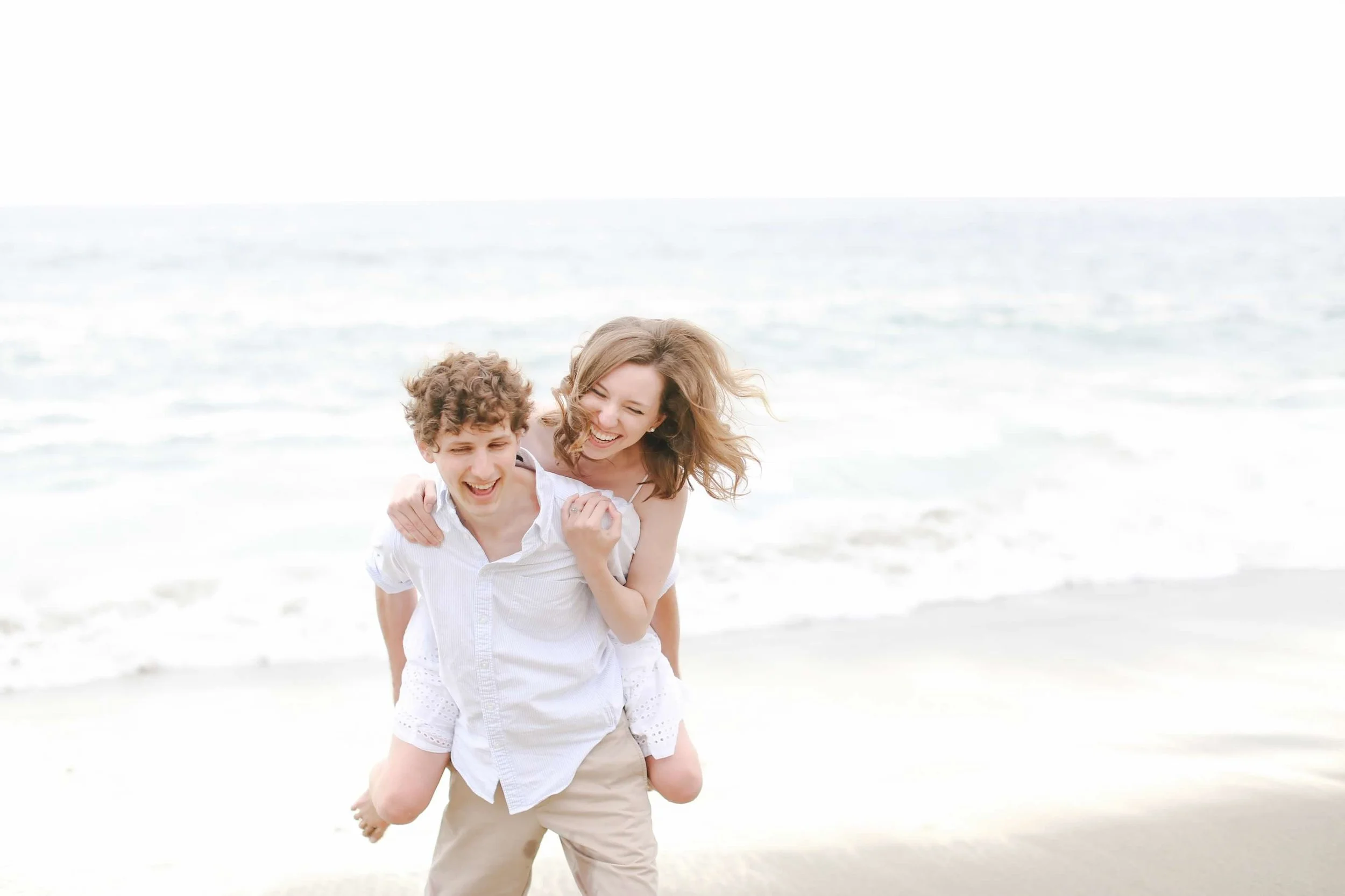 A happy man giving a piggyback ride to a young woman along the shoreline at the beach.