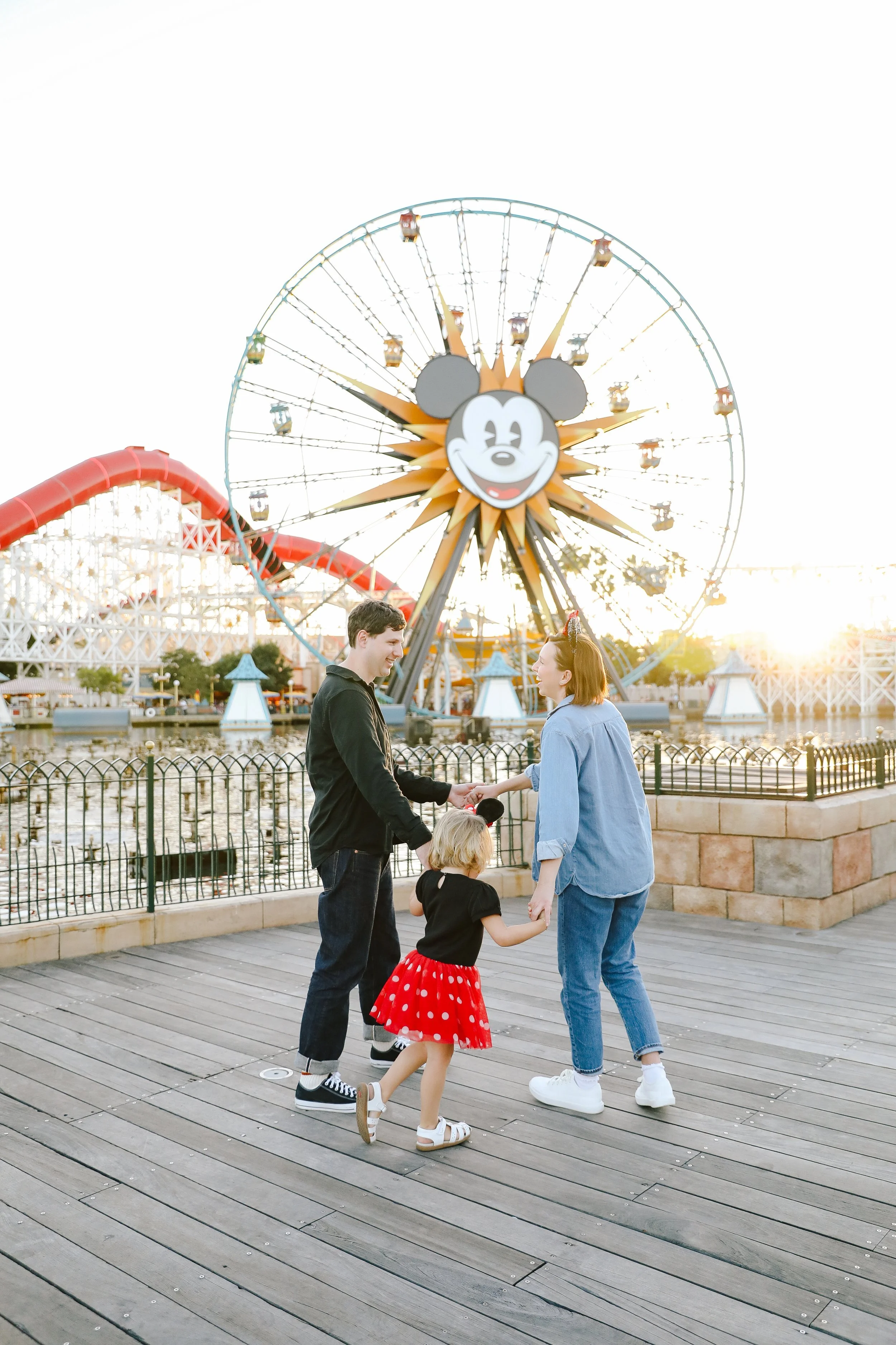 A family of three holding hands and playing on a wooden boardwalk at an amusement park with a large Mickey Mouse Ferris wheel in the background during sunset.