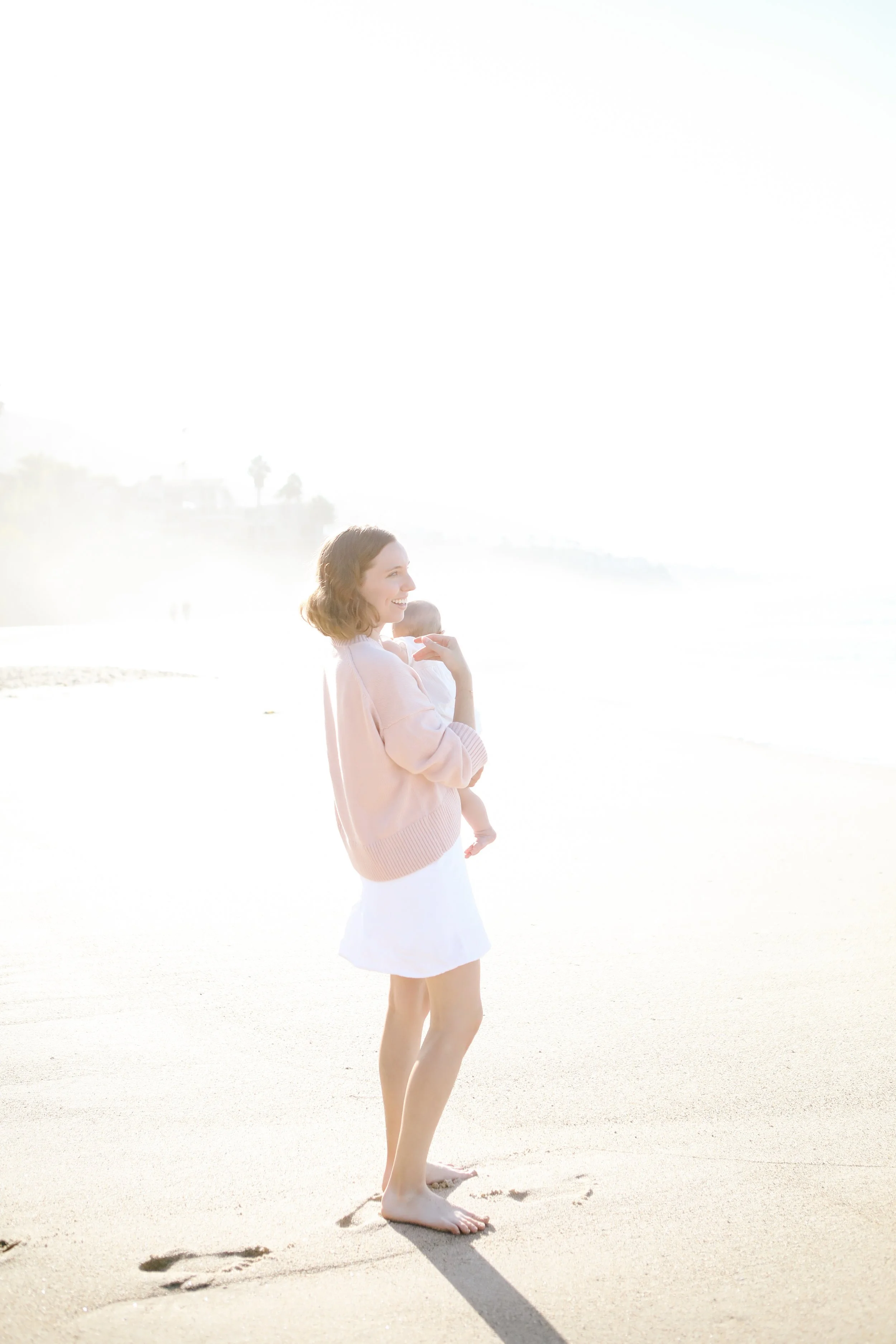 A woman in a pink sweater and light-colored skirt holding a baby on the beach. They are barefoot, with footprints in the sand, and looking out toward the ocean in bright sunlight.
