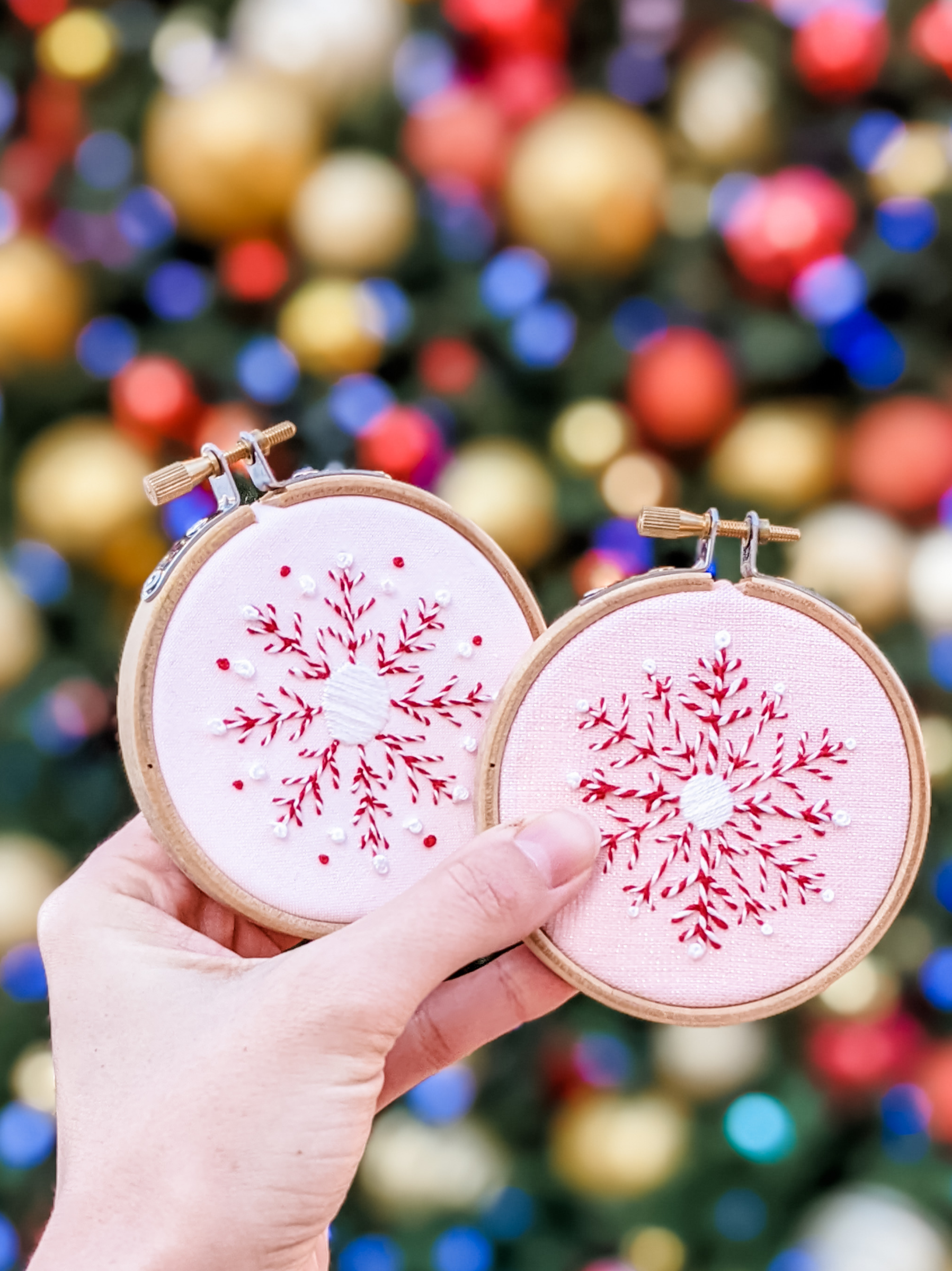 A person's hand holding two pink circular embroidery hoops with snowflake designs embroidered on pink fabric, in front of a blurred Christmas tree with colorful ornaments and lights.