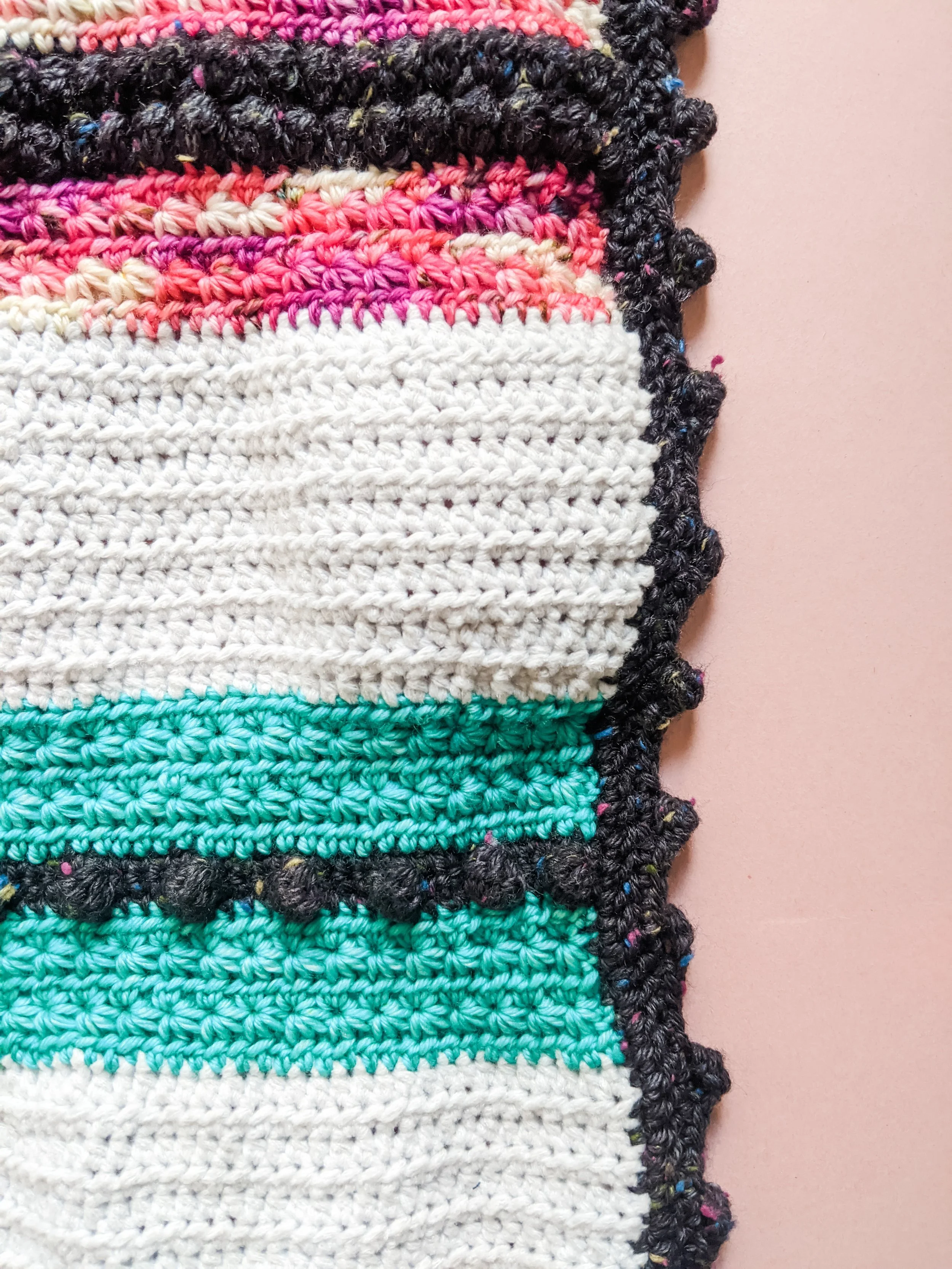 Close-up of a multicolored crocheted blanket with varying stripes including black, pink, white, turquoise, and multicolored yarn, laid on a pink surface.